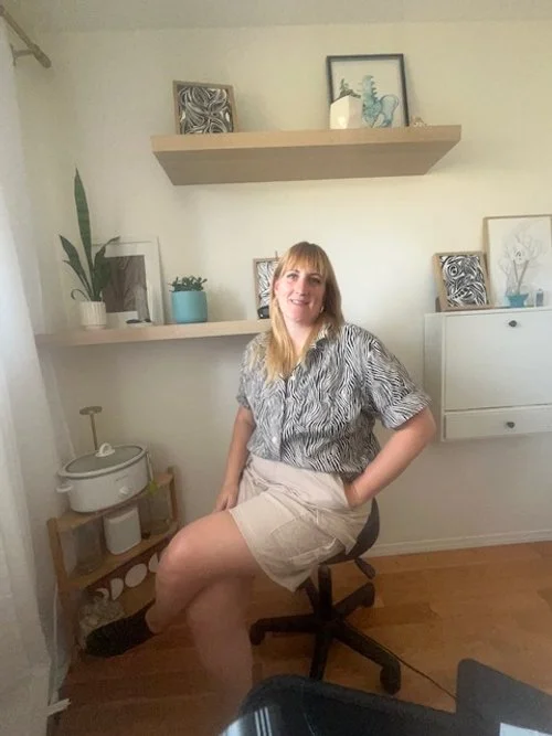 A woman with blond hair and bangs sitting on a black office chair in a room with white walls and wooden floors. She is smiling and wearing a patterned shirt and beige shorts. Behind her are white shelves with decorative items, including plants, framed artwork, and a small white cabinet.