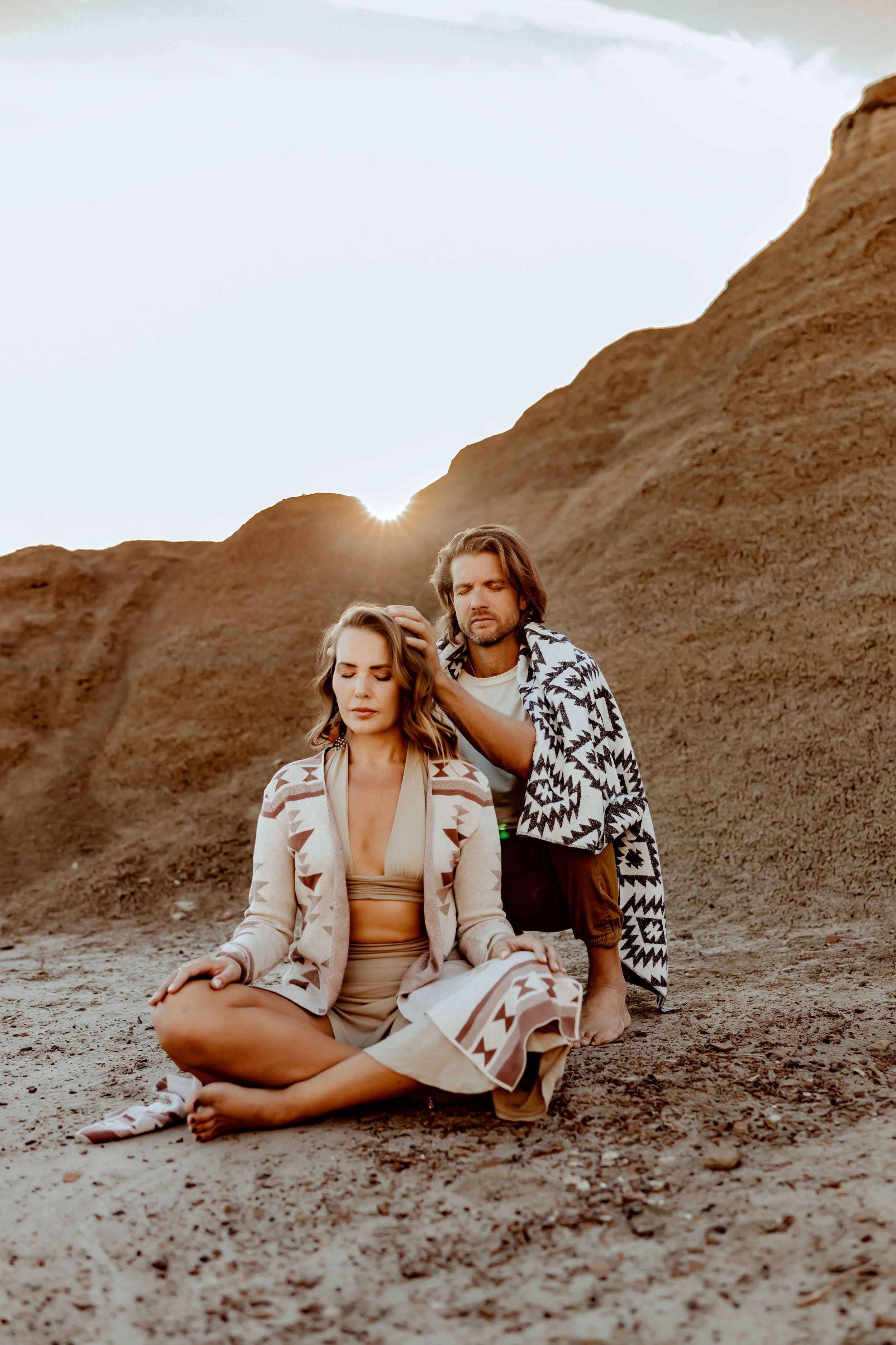 A woman sitting cross-legged on the ground with eyes closed, a man kneeling beside her touching her head with his hand, both dressed in patterned clothing, in a desert-like landscape with cliffs and a setting sun.