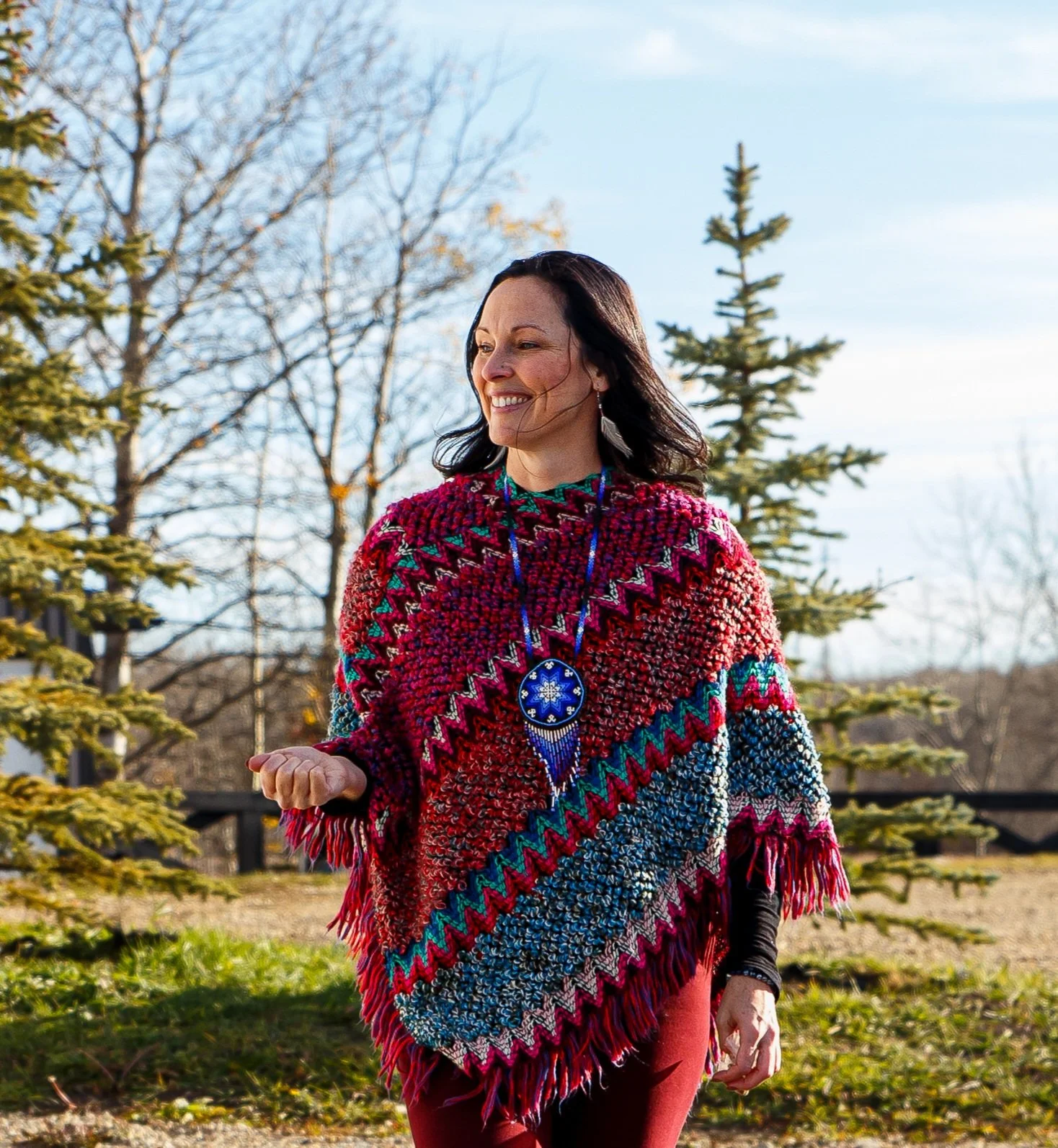 A woman with black hair smiling outdoors, wearing a colorful, knit poncho with zigzag patterns in multiple colors and a large blue beaded necklace, standing in front of pines and bare trees during daytime.