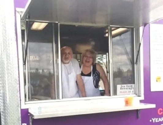 Two smiling people working at an ice cold beverage stand, wearing aprons, with a purple exterior and a small orange cup on the counter.