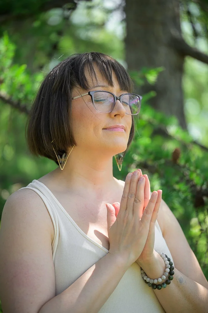 A woman with short dark hair, glasses, and earrings, practicing yoga outdoors with her hands in prayer position, surrounded by green trees.