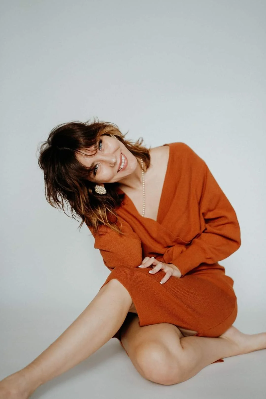 Woman with wavy brown hair smiling, sitting on the floor in a rust-colored dress, against a plain light background.