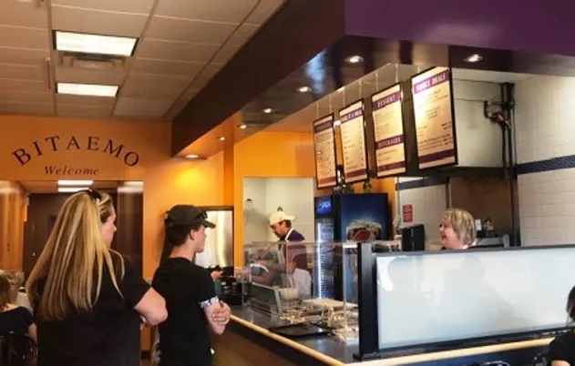 Customers order at the counter of a restaurant named Bitaemo with a menu displayed overhead and staff serving food behind a glass partition.