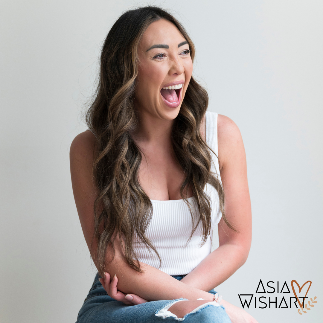 A woman with long wavy brown hair laughing with her mouth open, wearing a white sleeveless top and ripped jeans, sitting against a plain white background.