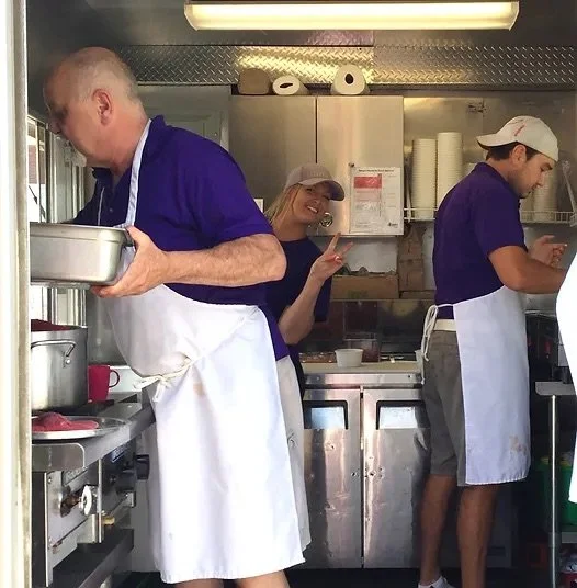 Three people working in a commercial kitchen, two men and a woman, all wearing aprons and hats. The woman is smiling and making a peace sign with her fingers.