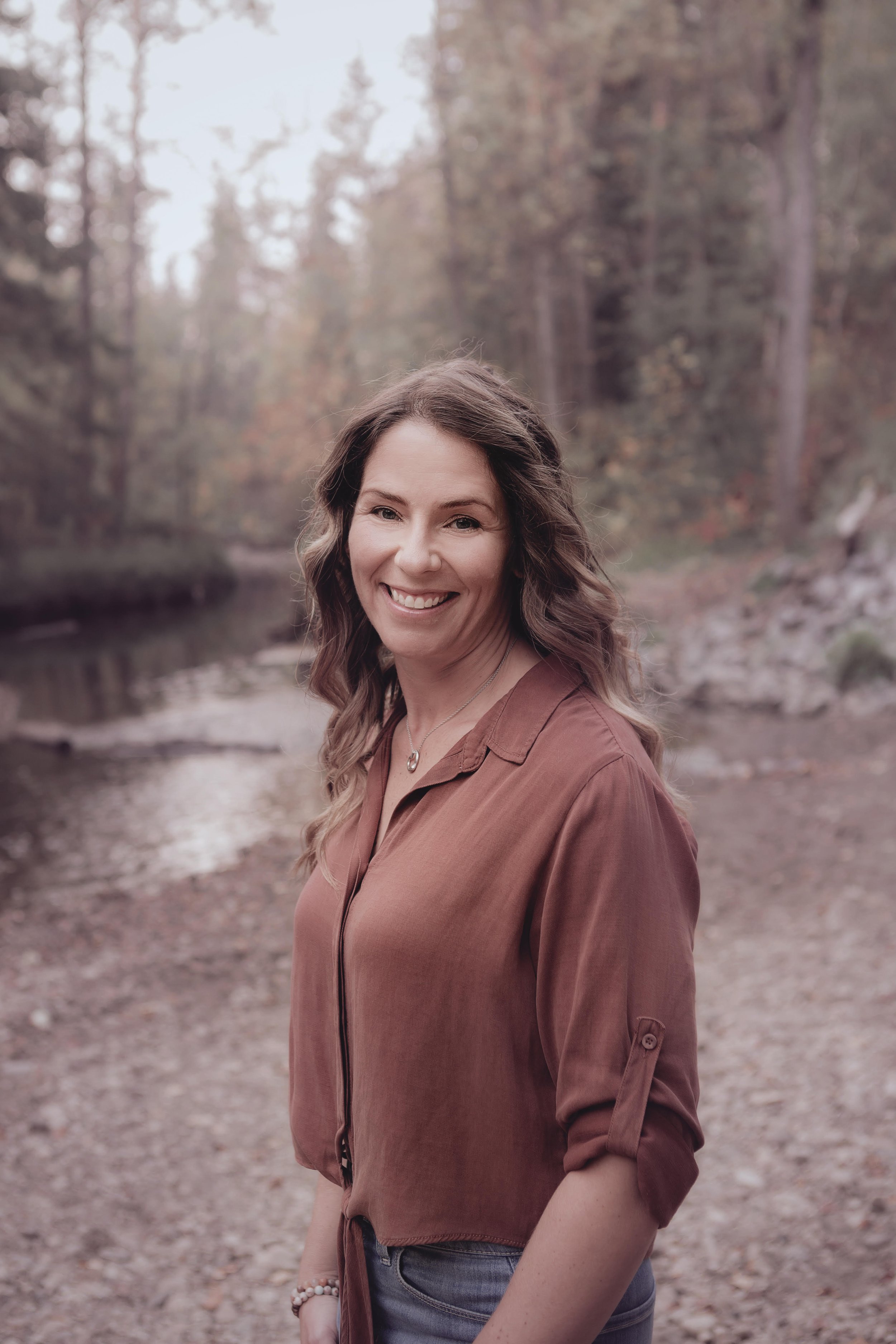 A smiling woman with wavy brown hair wearing a rust-colored blouse and jeans stands outdoors near a river and trees in autumn.