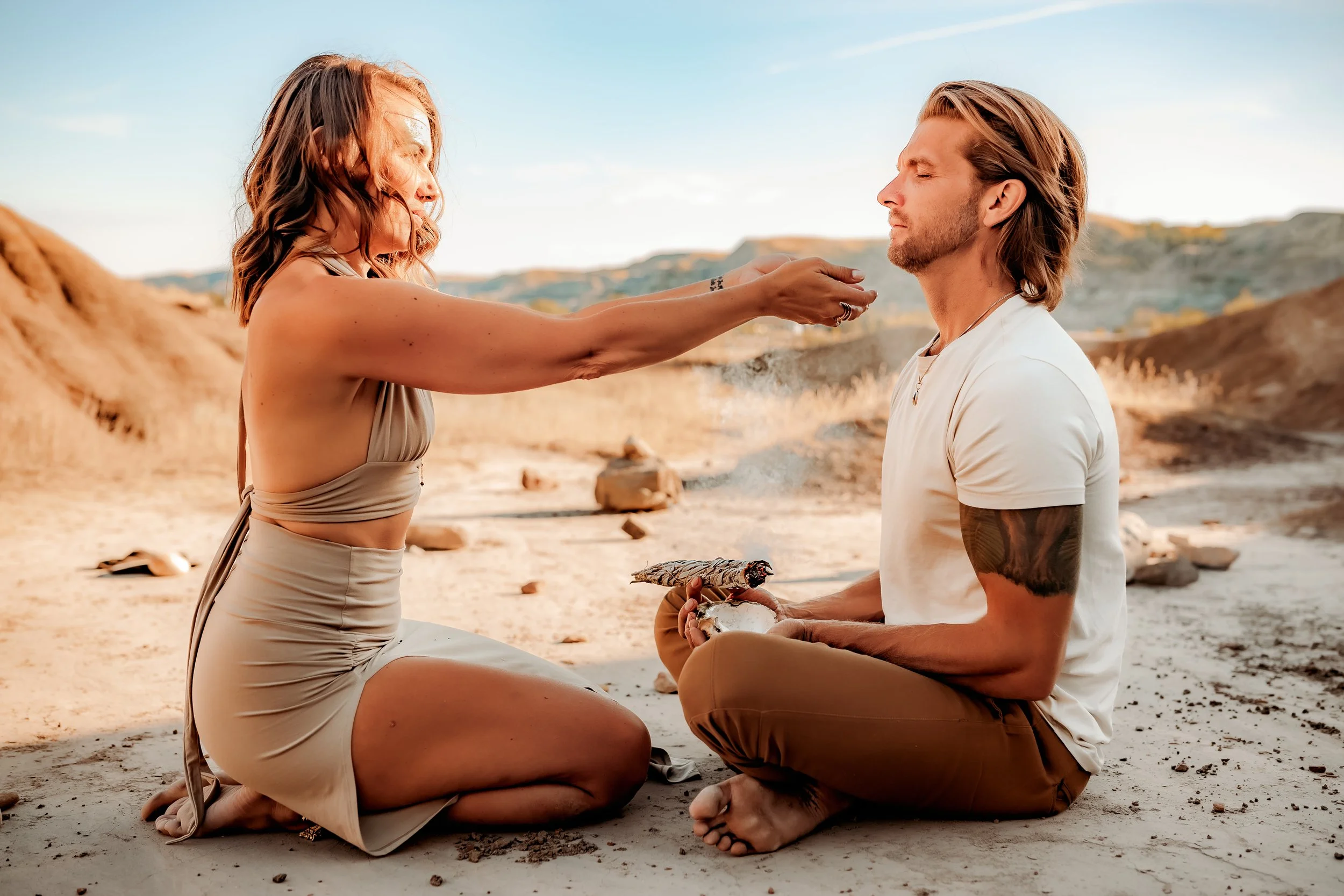 A woman is performing a spiritual or ceremonial ritual with a man in an outdoor desert landscape, with both sitting on the sandy ground.