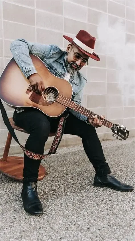 A man wearing a red hat, sunglasses, and a denim jacket playing an acoustic guitar while sitting on a wooden chair against a light-colored wall.