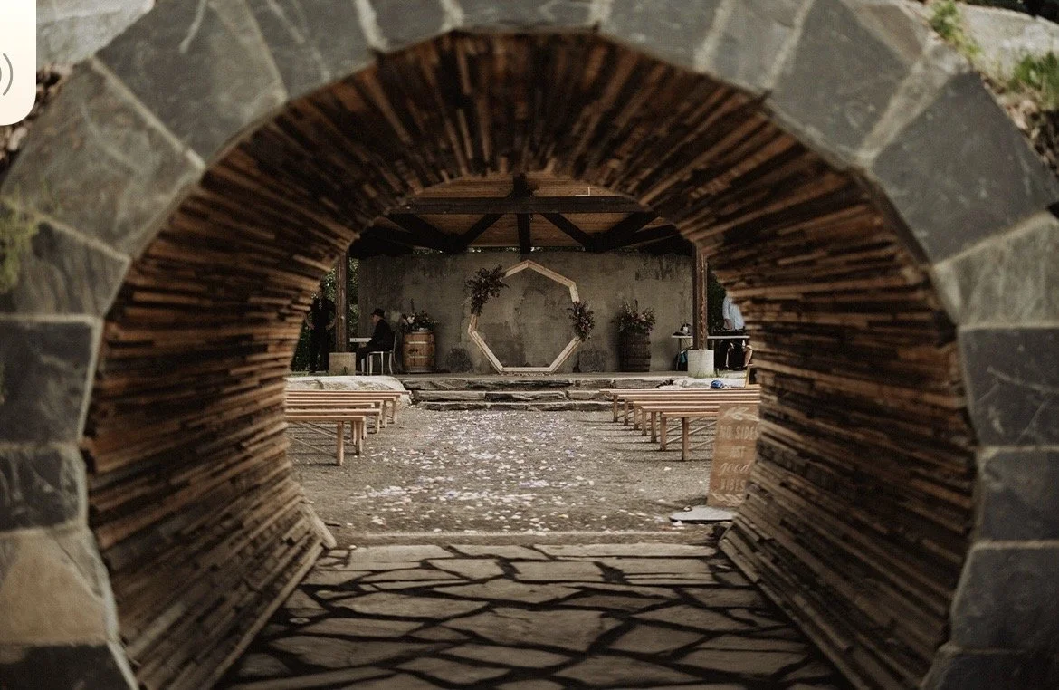 View through a circular stone tunnel leading to a rustic wedding or event setup with benches, flower arrangements, and a hexagonal floral display on a concrete wall.