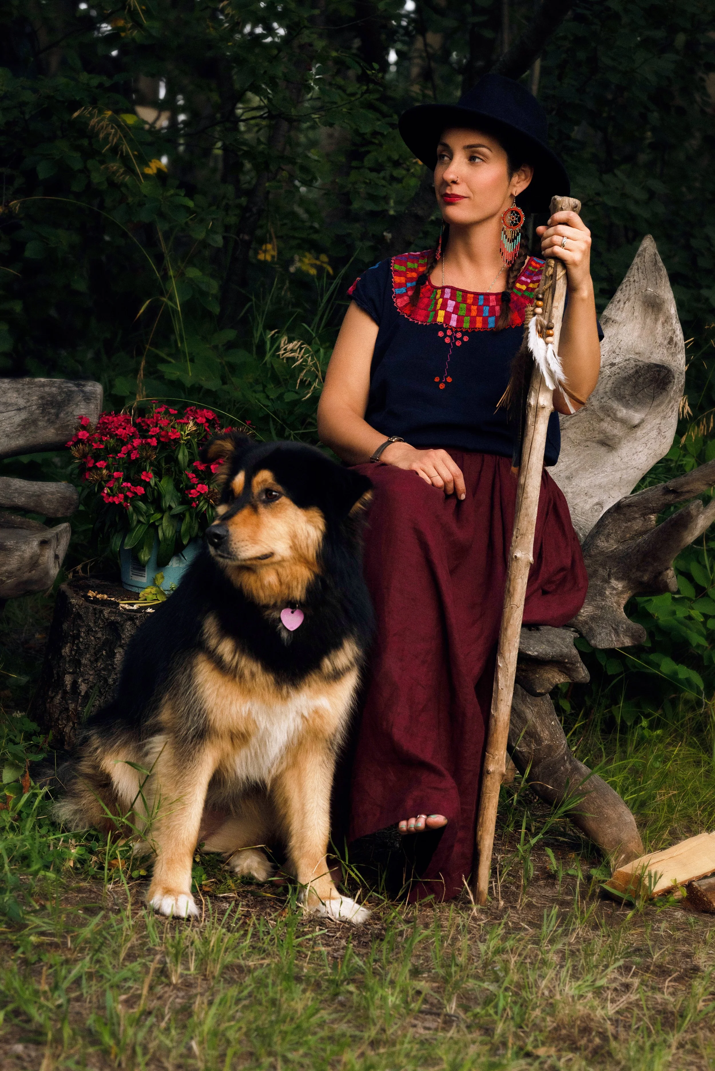 A woman dressed in traditional indigenous attire, sitting outdoors in a natural setting with a dog and flowers nearby.