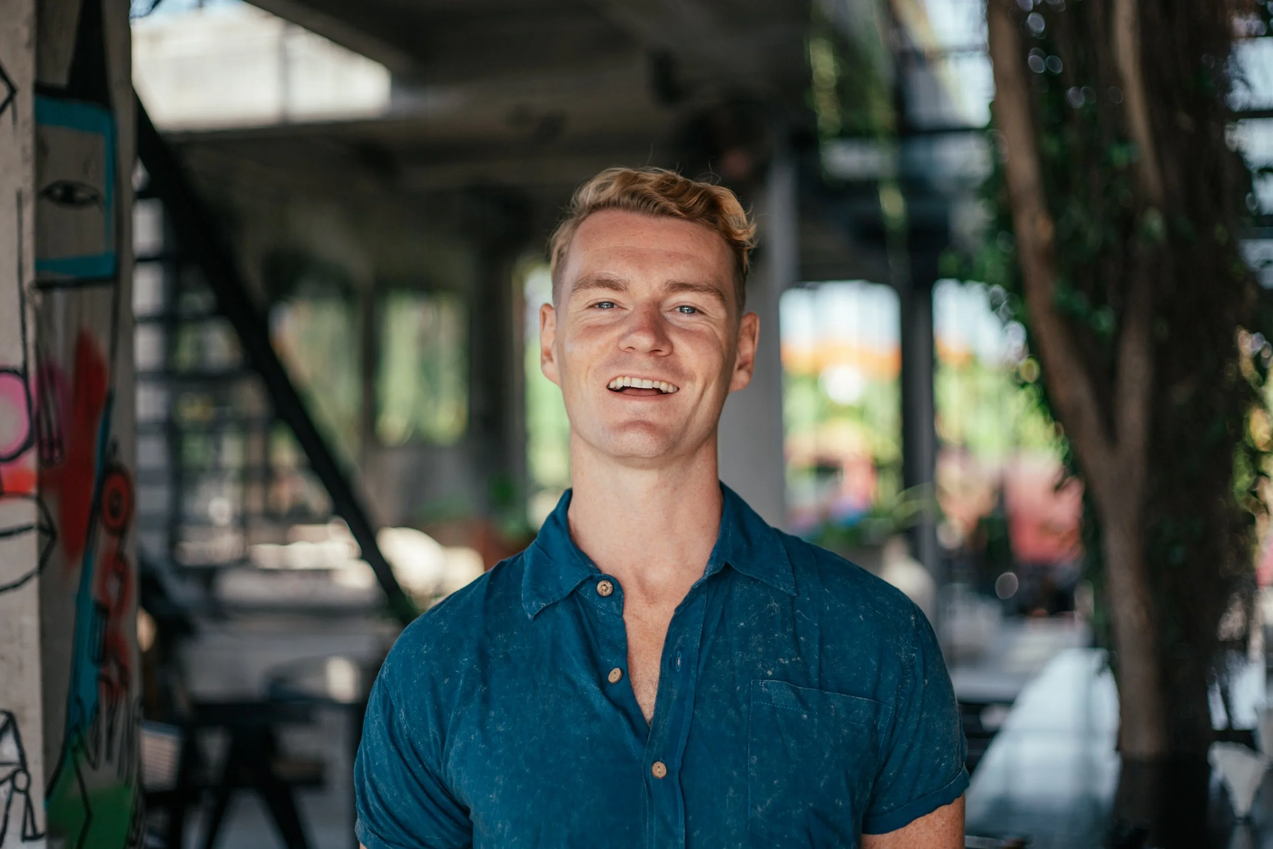 A smiling young man with short blond hair wearing a blue shirt, standing in an outdoor setting with blurred background of trees and structures.