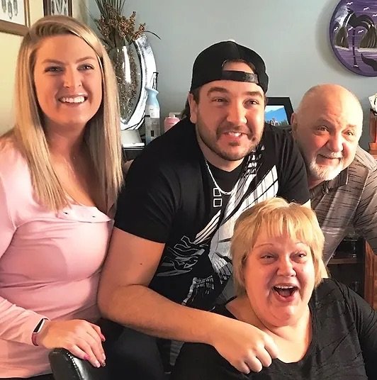 Four smiling people gathered together indoors, including a woman in a pink shirt on the left, a man in a black cap and T-shirt in the center, an older man with a bald head and beard on the right, and a woman with short blonde hair in front, all close to each other.