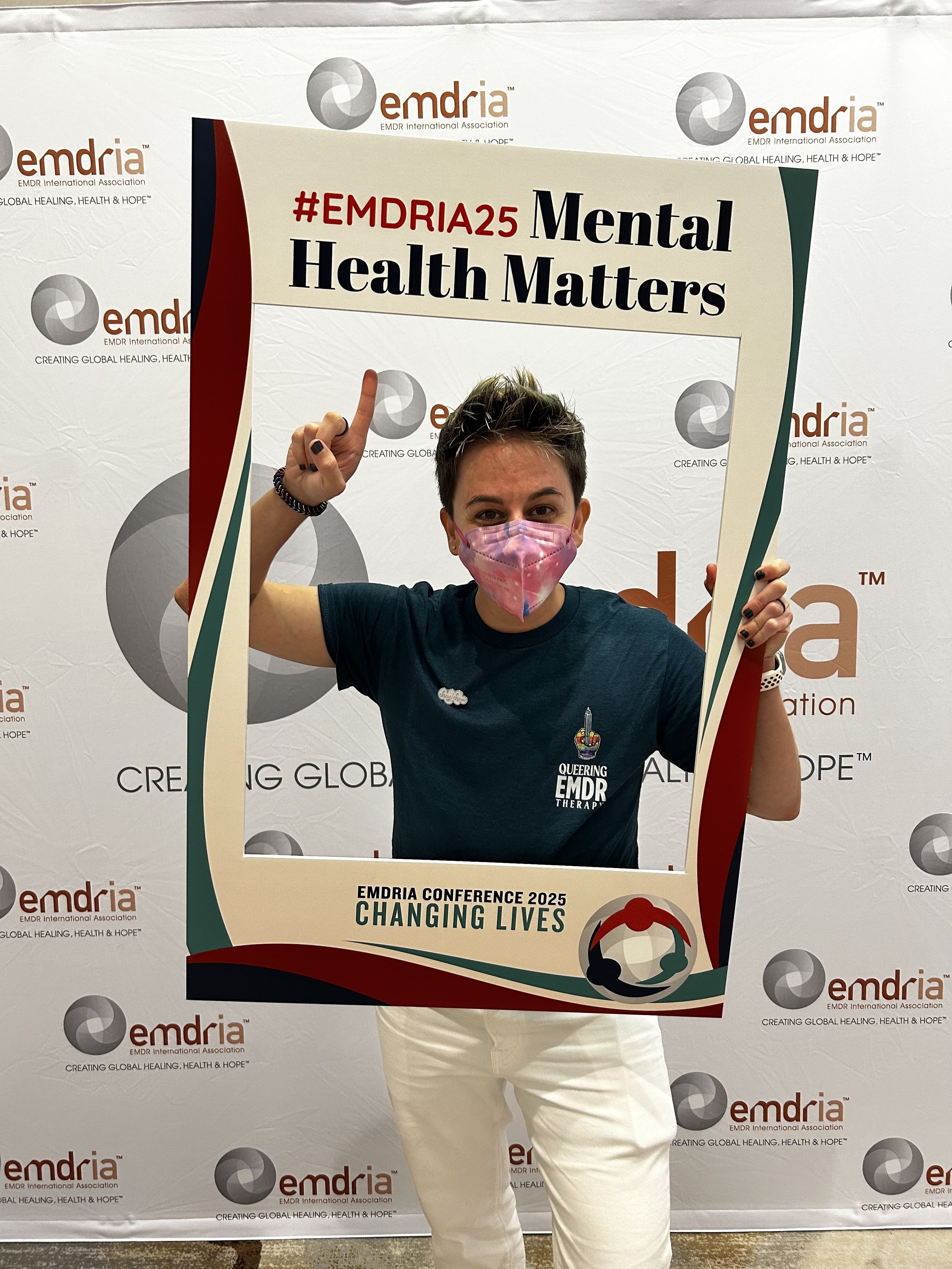 Woman wearing a pink mask holding a large sign that says '#EMDRIA25 Mental Health Matters' at an EMDR conference.