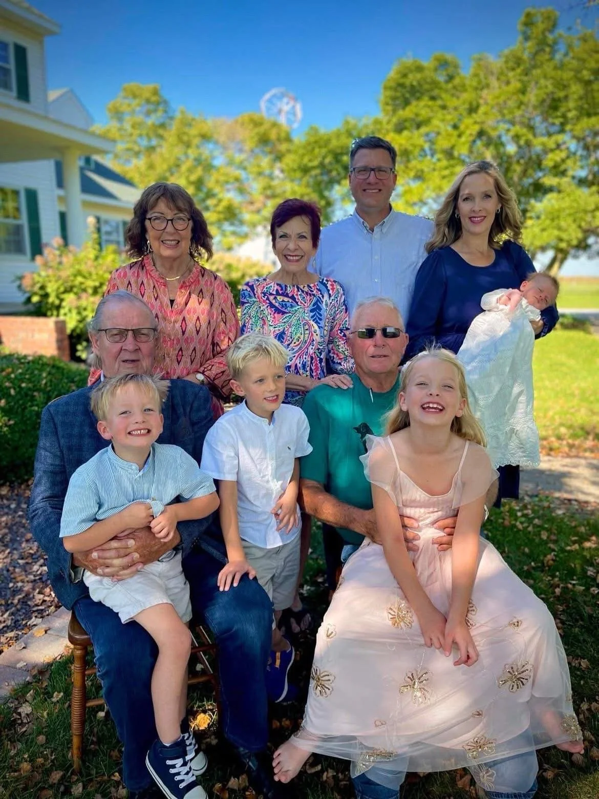 Family photo outdoors on a sunny day with multiple generations, including children, adults, and seniors, smiling in front of a house and trees.