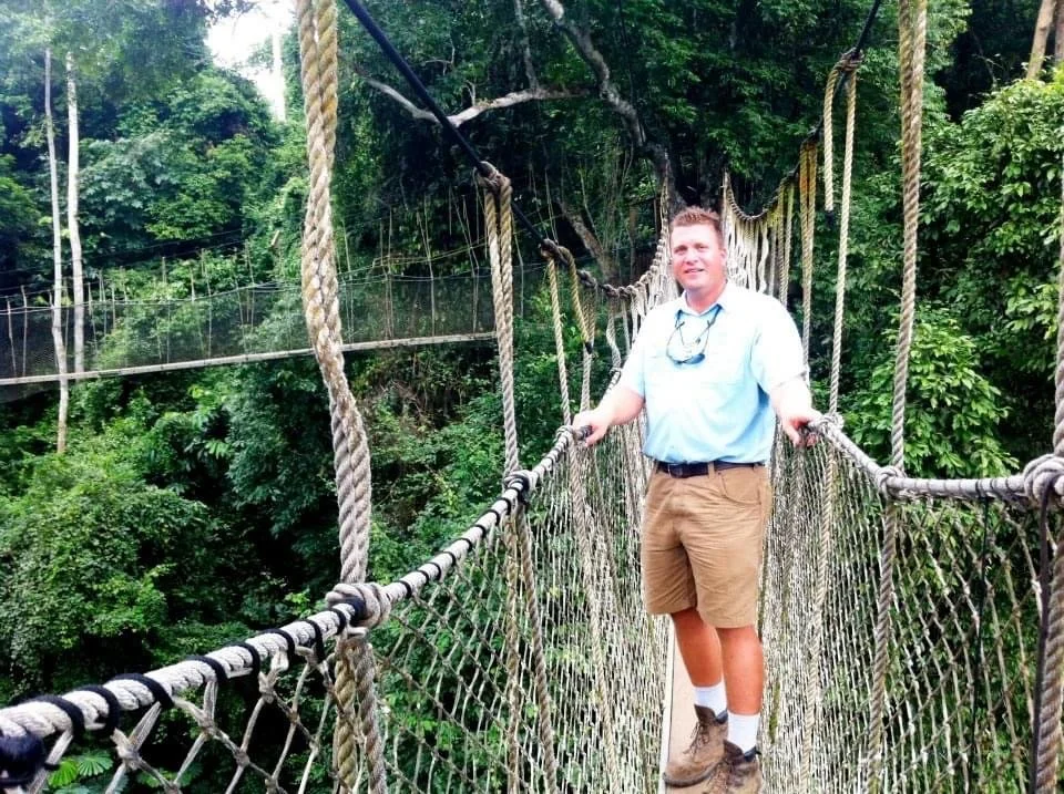 A young man stands on a narrow rope bridge in a lush green forest, holding onto the ropes on either side, with trees and greenery in the background.