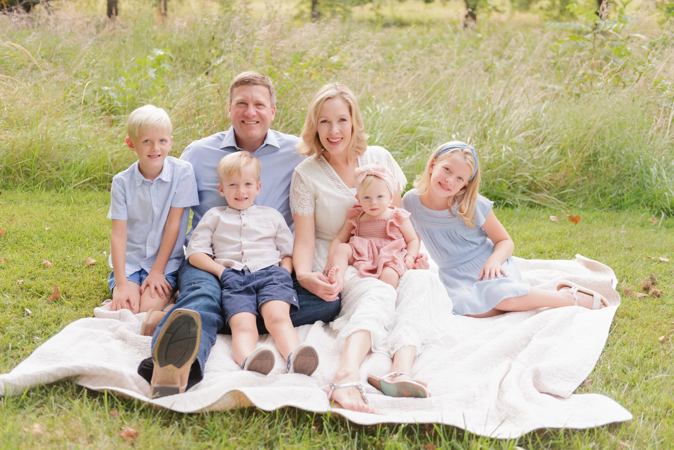 A happy family of seven sitting on a blanket on the grass in a park, surrounded by greenery and tall grass.