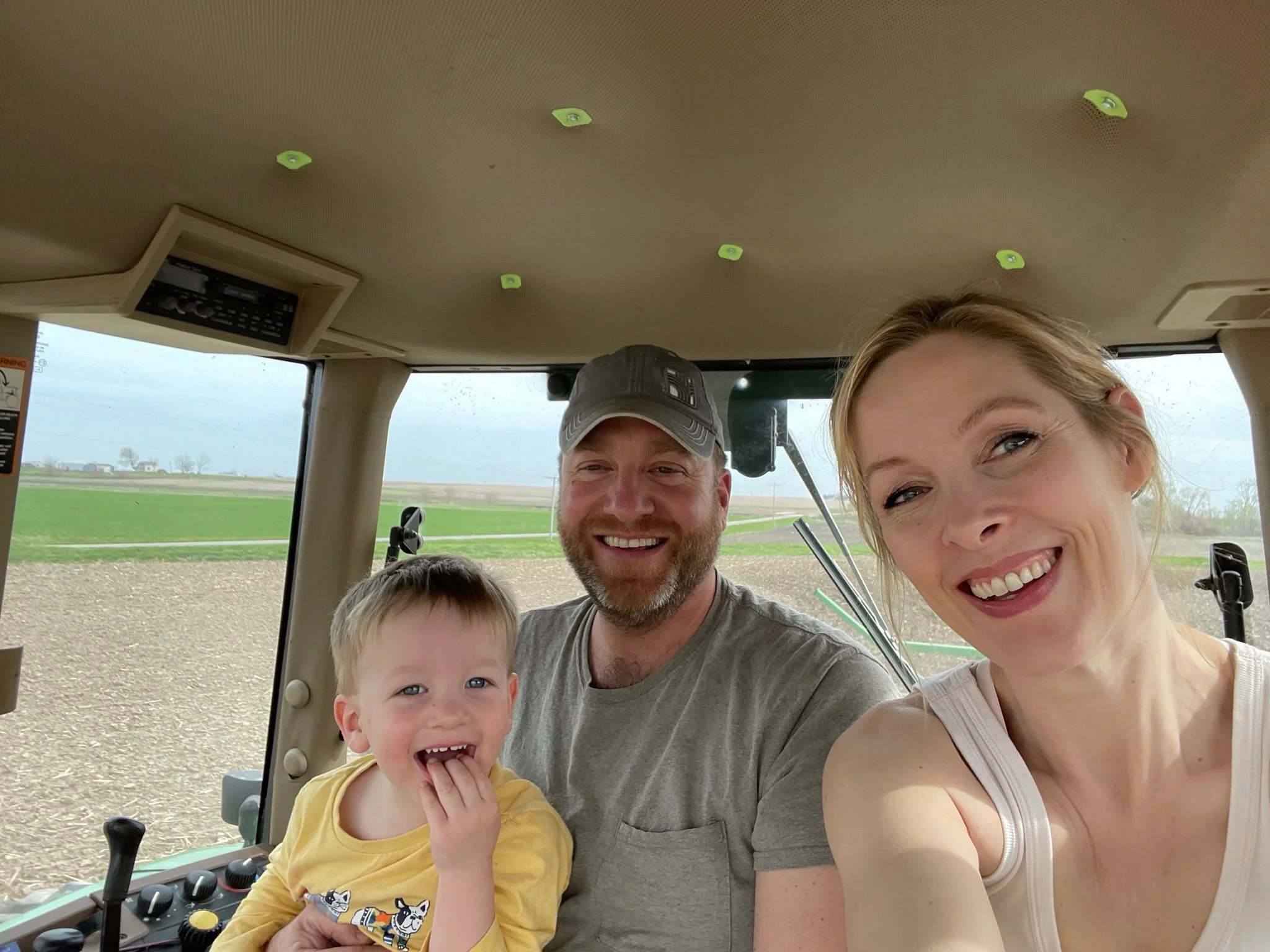 A smiling family of three inside a vehicle, with two adults and a young child, in front of a green rural landscape.