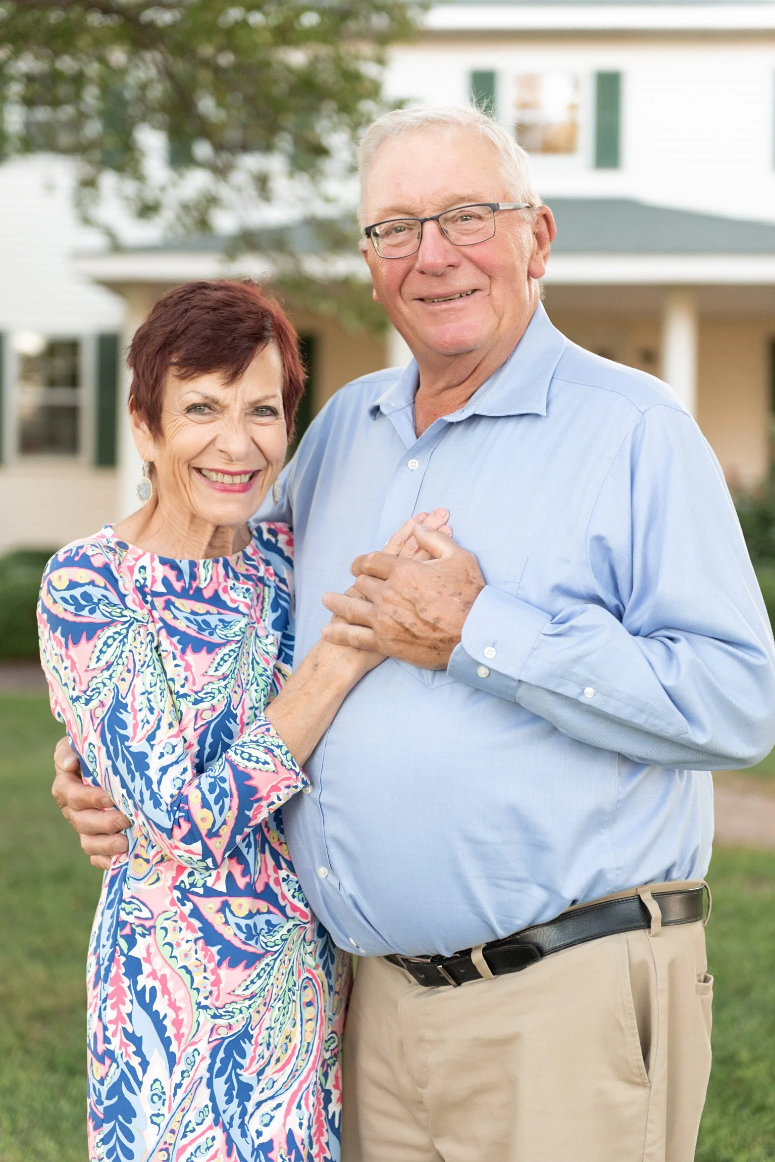An elderly couple dancing outdoors with a house in the background. The woman has short dark hair and is wearing a colorful patterned dress, and the man has gray hair, glasses, and is wearing a light blue button-down shirt and beige pants.