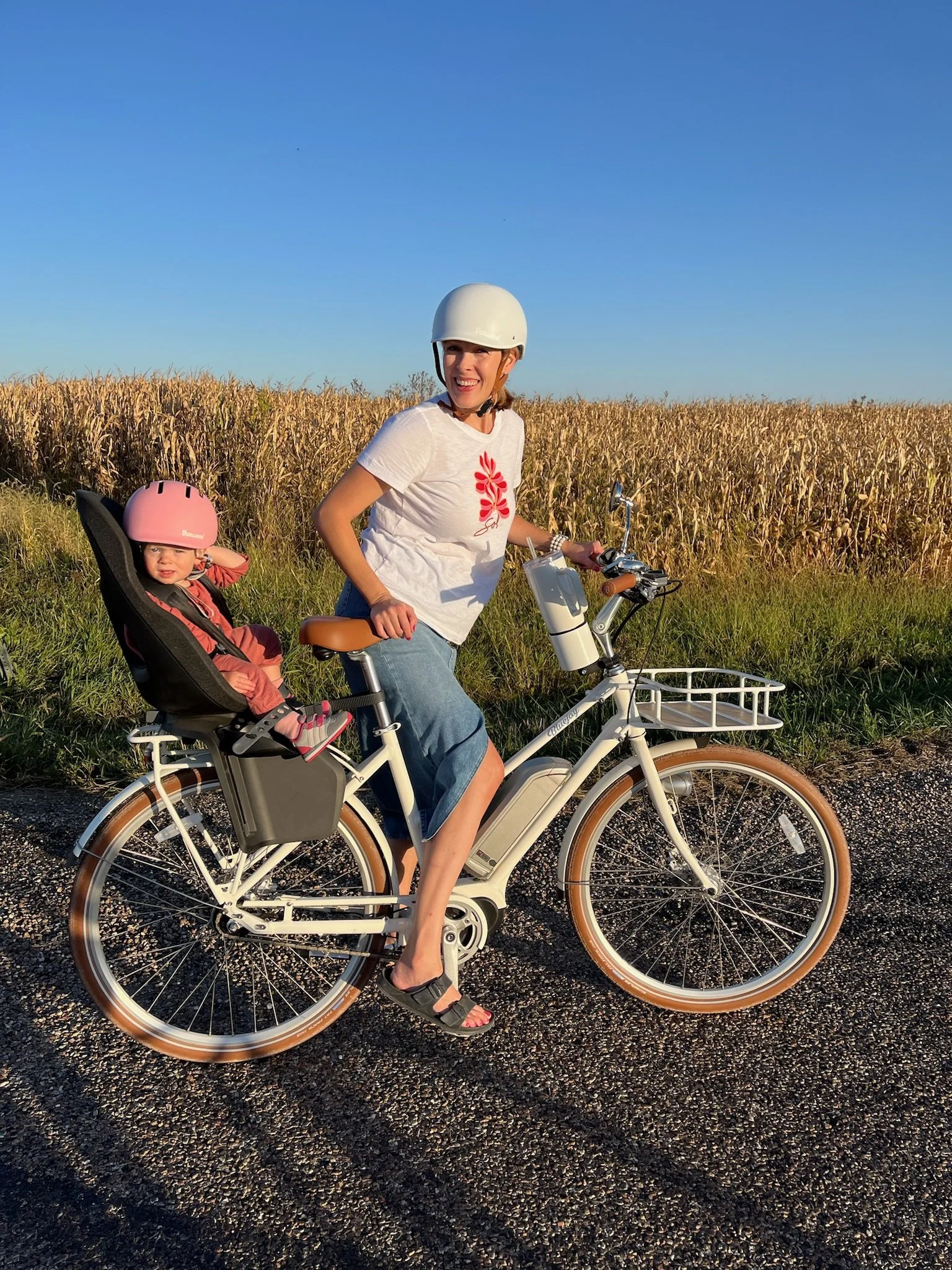 Woman riding a white bicycle with a child in a child seat in the back, both wearing helmets, on a rural road with a field of tall grass or crops and a clear blue sky in the background.