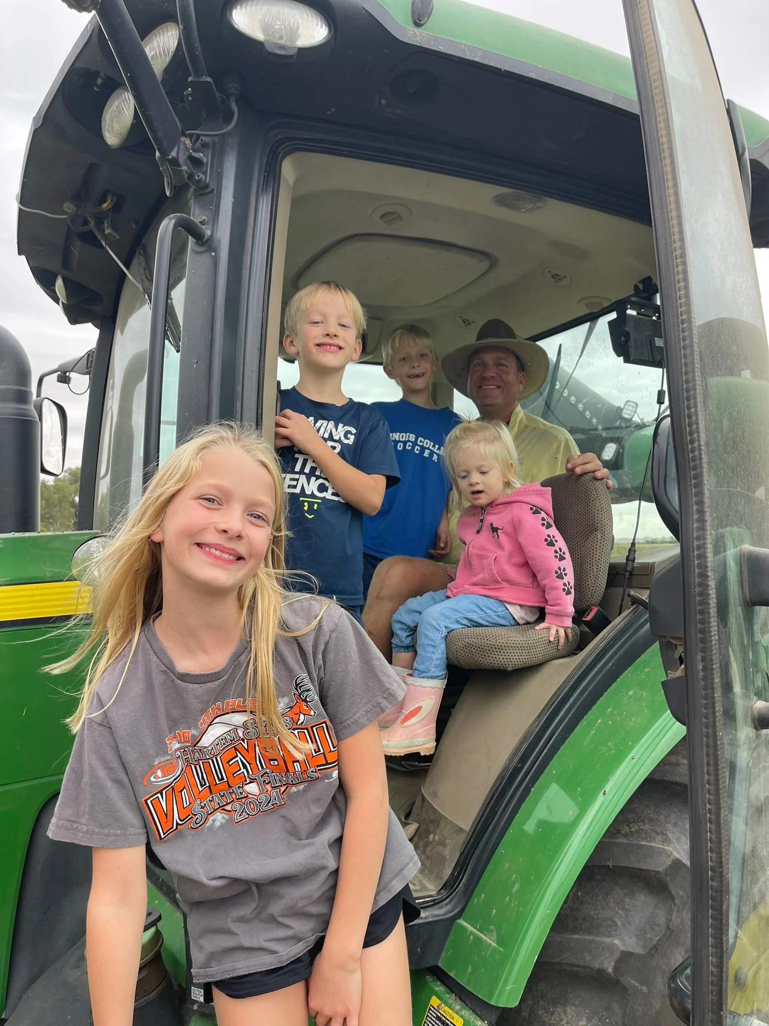 A young girl with long blonde hair smiling in front of a green tractor with multiple children and a man sitting inside.