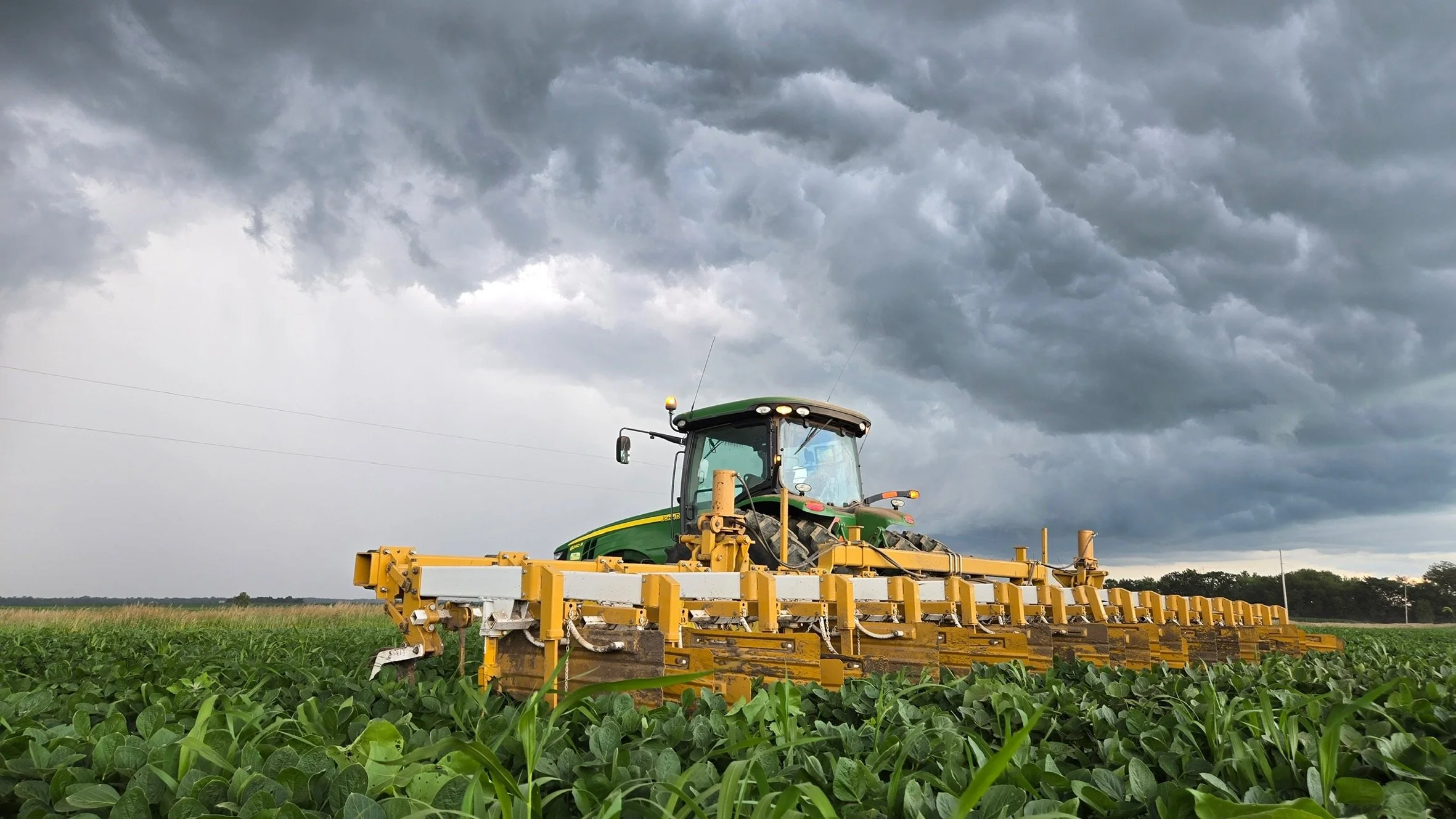 A green tractor with yellow equipment working in a field of green crops under a cloudy sky.