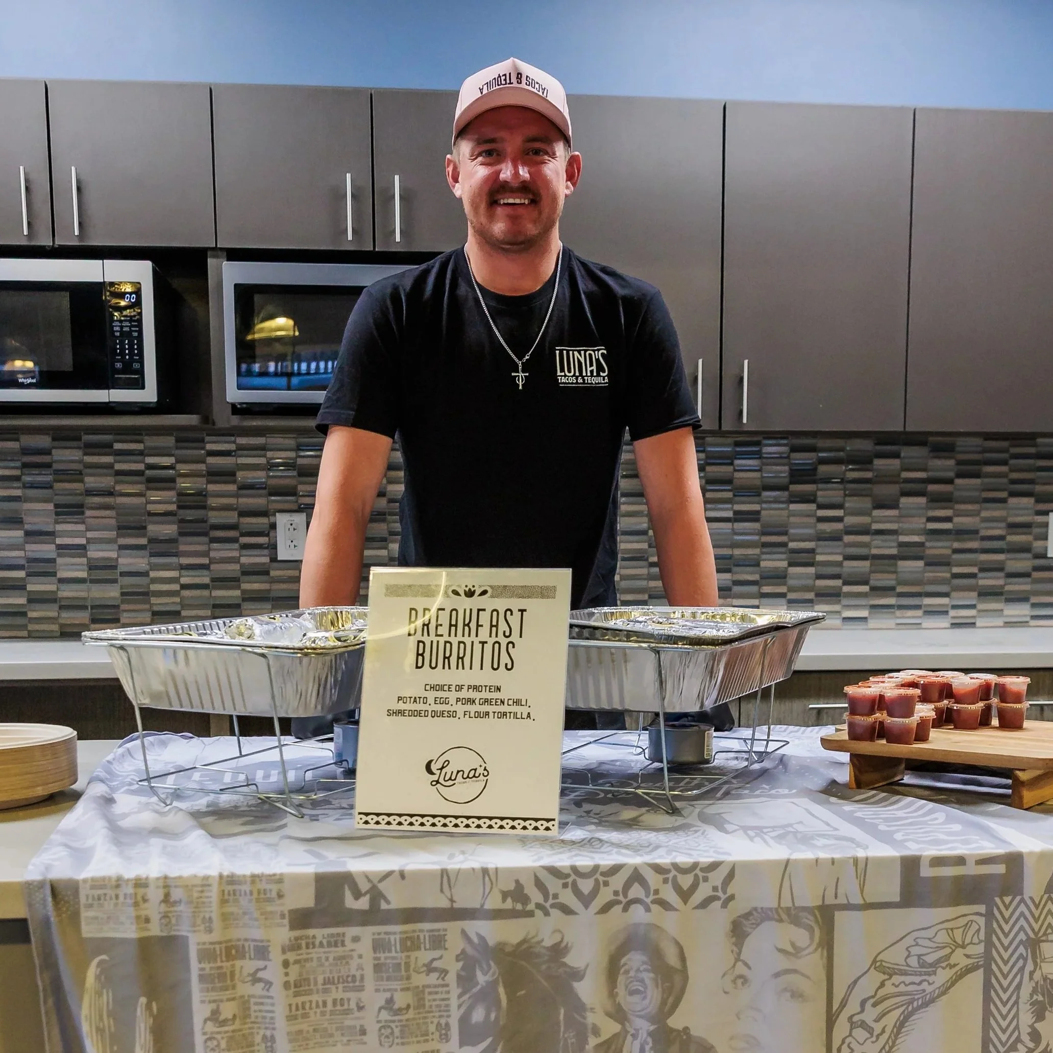 A smiling man stands behind a Luna’s Tacos and Tequila breakfast burrito setup with catering trays and salsa cups in a breakroom kitchen.
