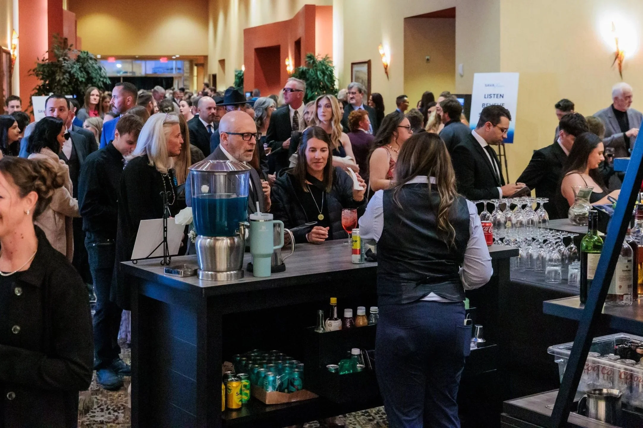 A large crowd mingles in a warmly lit event lobby during a formal reception, with guests gathered around a busy bar and talking in small groups. The space feels lively and elegant, with many attendees dressed in cocktail or evening attire.