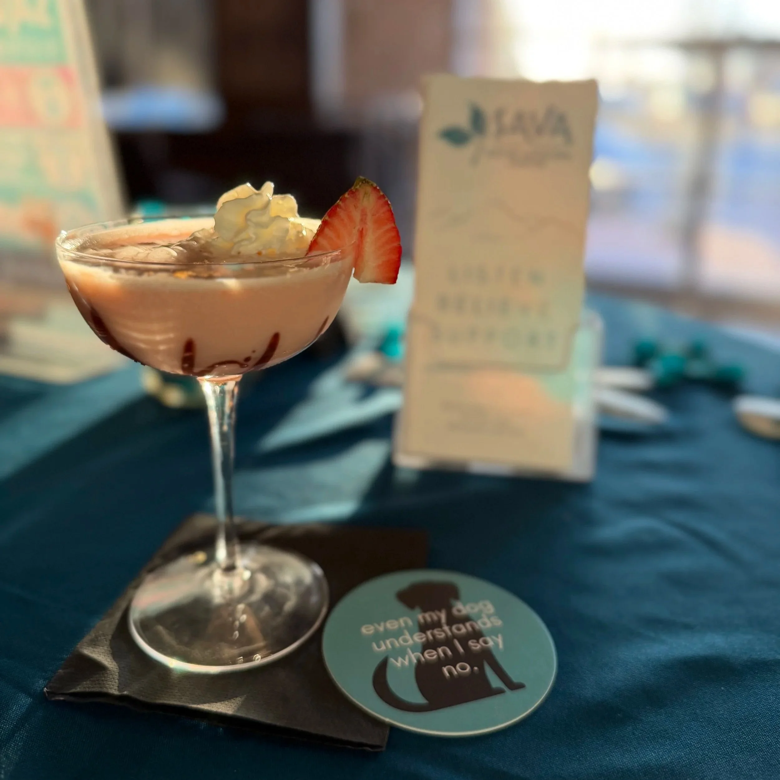 Pink cocktail with whipped cream and a strawberry slice on a teal tablecloth beside SAVA materials and a teal coaster.