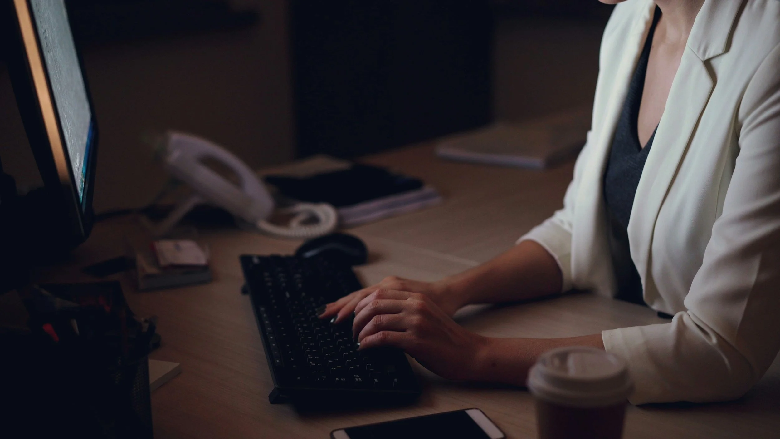A person in a white blazer types at a computer desk in a dimly lit office. A monitor, keyboard, phone, and takeaway coffee cup are visible, creating a focused workplace scene.
