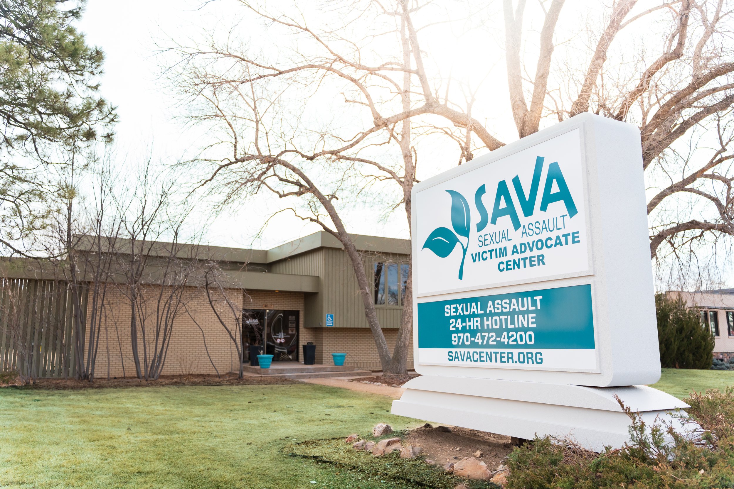 Daytime exterior of The SAVA Center building, featuring a large freestanding sign in the foreground with the organization’s name, sexual assault hotline number, and website, surrounded by lawn, trees, and the office building behind it.