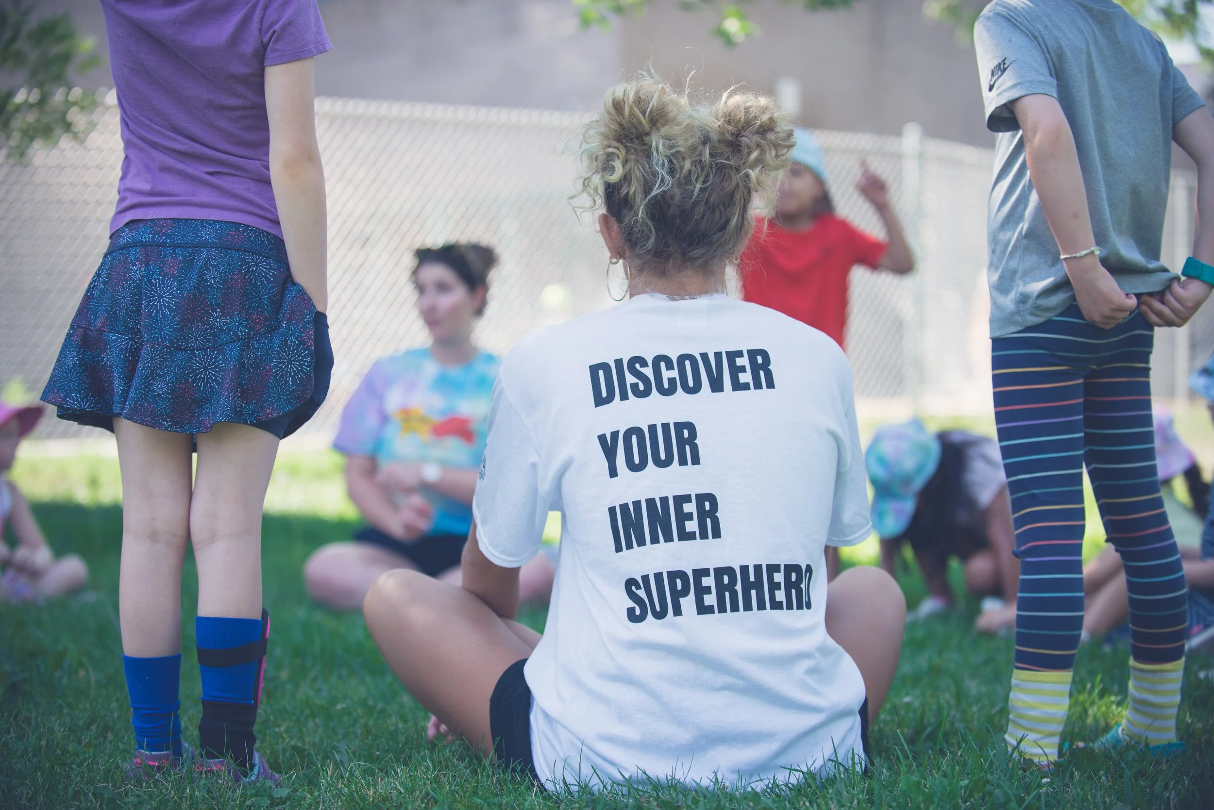 A group of children and adults sit outdoors on grass during an activity, with one person wearing a shirt that says ‘Discover Your Inner Superhero.’