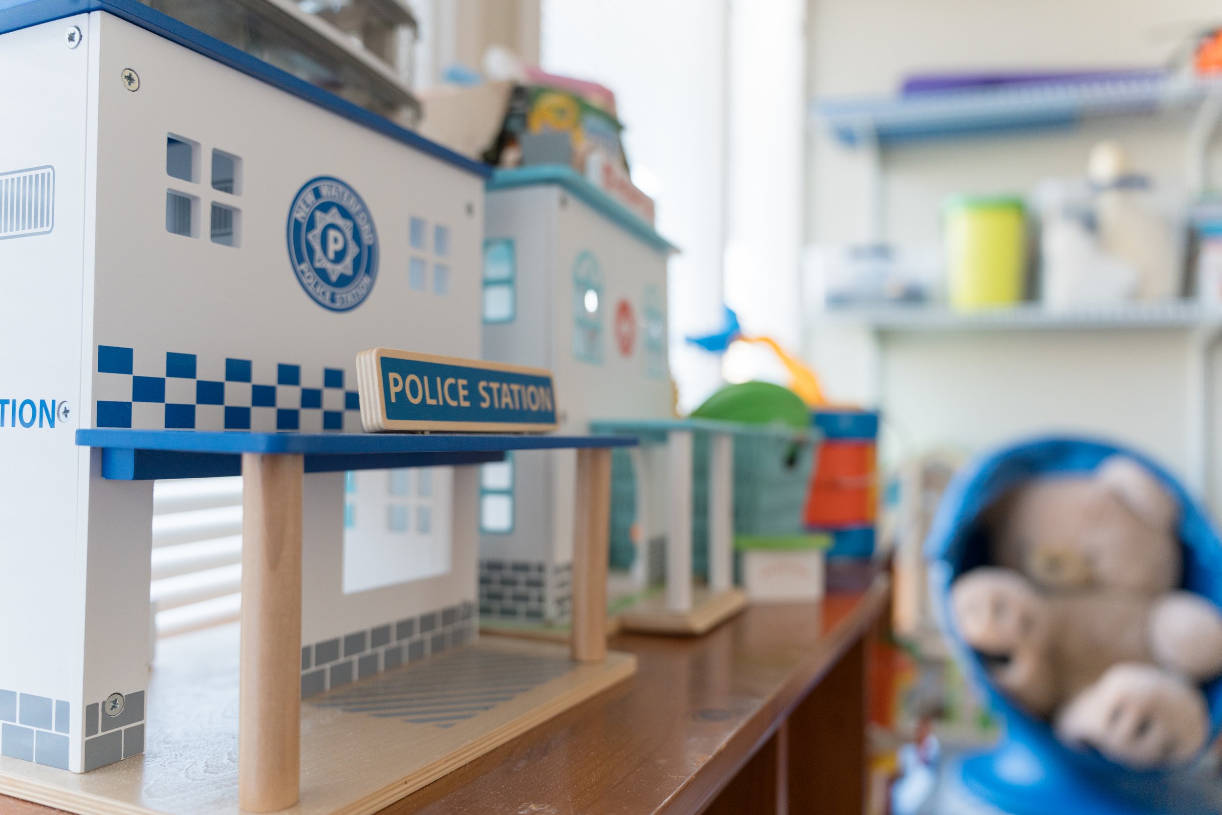 Close-up of a wooden toy police station on a shelf in a children’s playroom, with other toy buildings, colorful supplies, and a stuffed animal softly out of focus in the background.