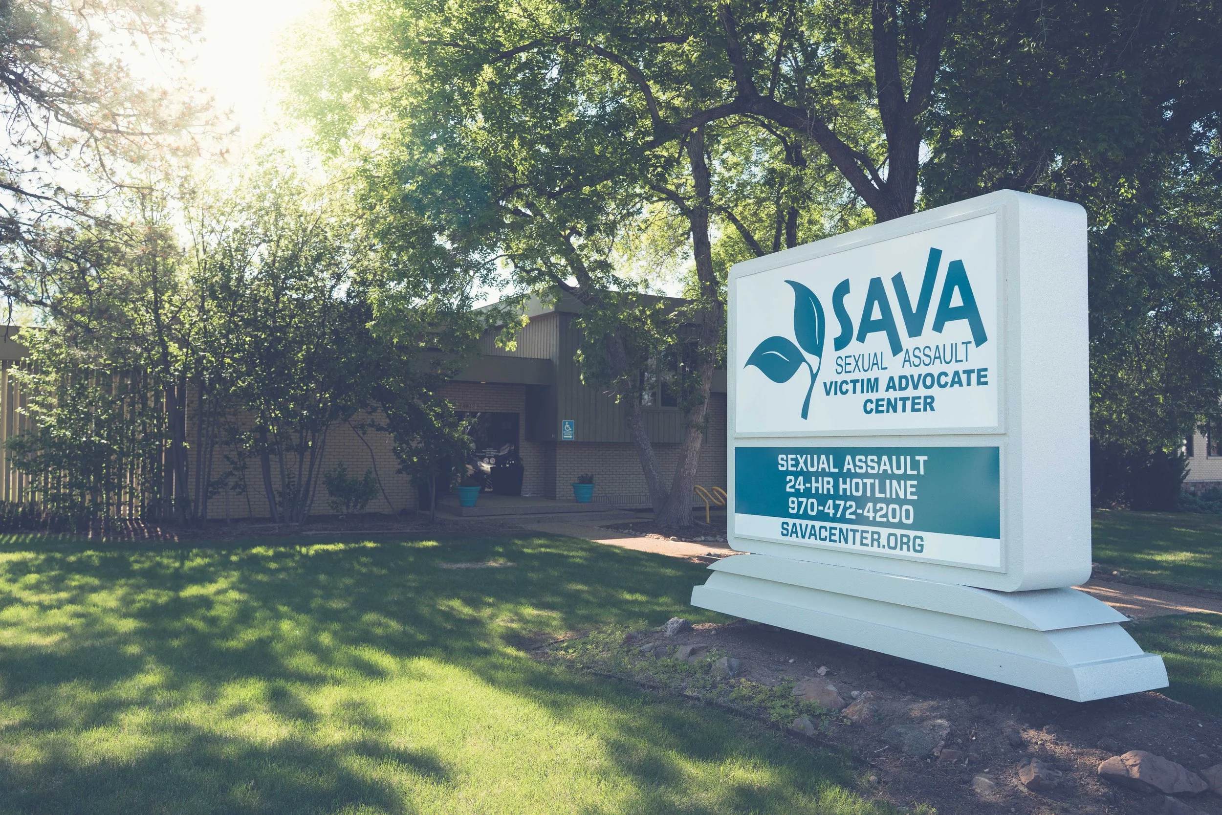 Daytime exterior of The SAVA Center building, featuring a large freestanding sign in the foreground with the organization’s name, sexual assault hotline number, and website, surrounded by lawn, trees, and the office building behind it.