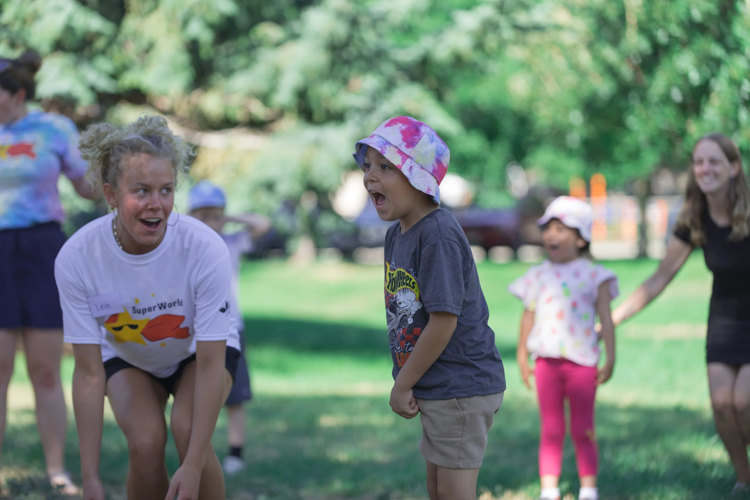 Children and adults play together on a grassy lawn, with one child in a pink bucket hat smiling with excitement in the foreground.