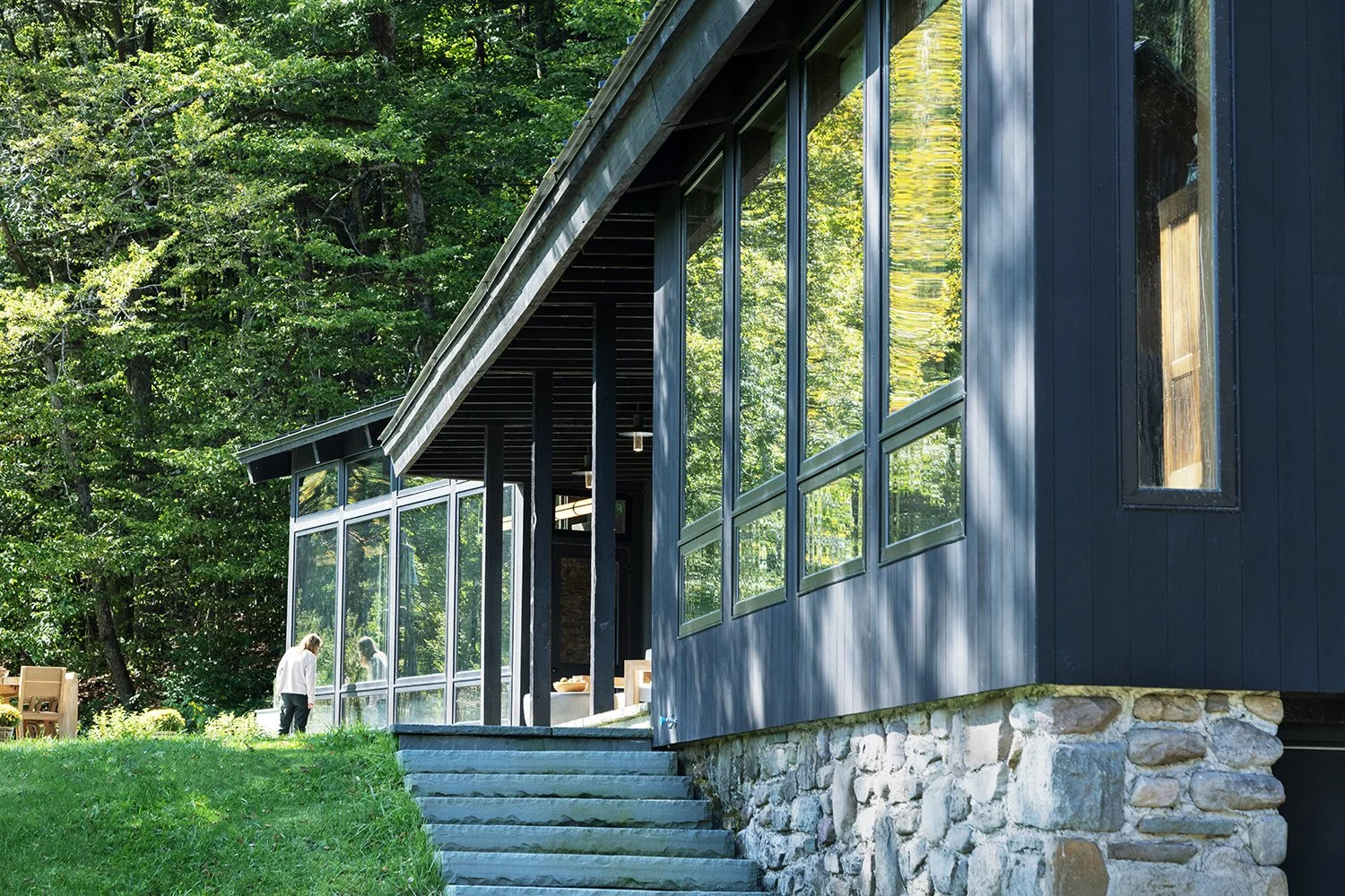 A modern house with a stone foundation, dark blue siding, and large glass windows, surrounded by green trees and grass. A person is standing outside on the steps.