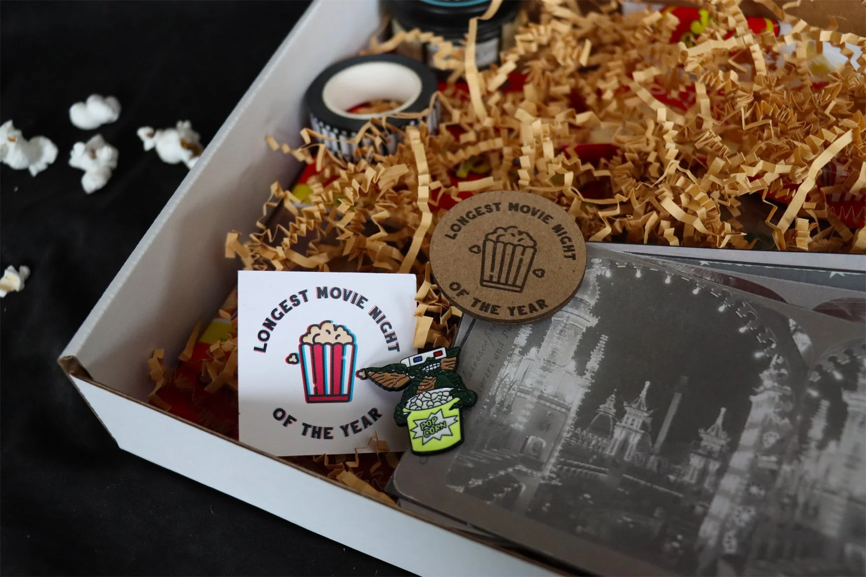 Close-up of gift box showing small card with cyan-and-red "3D" popcorn logo, enamel pin of Gremlin wearing 3D glasses and holding popcorn bucket, photo of brightly lit building.