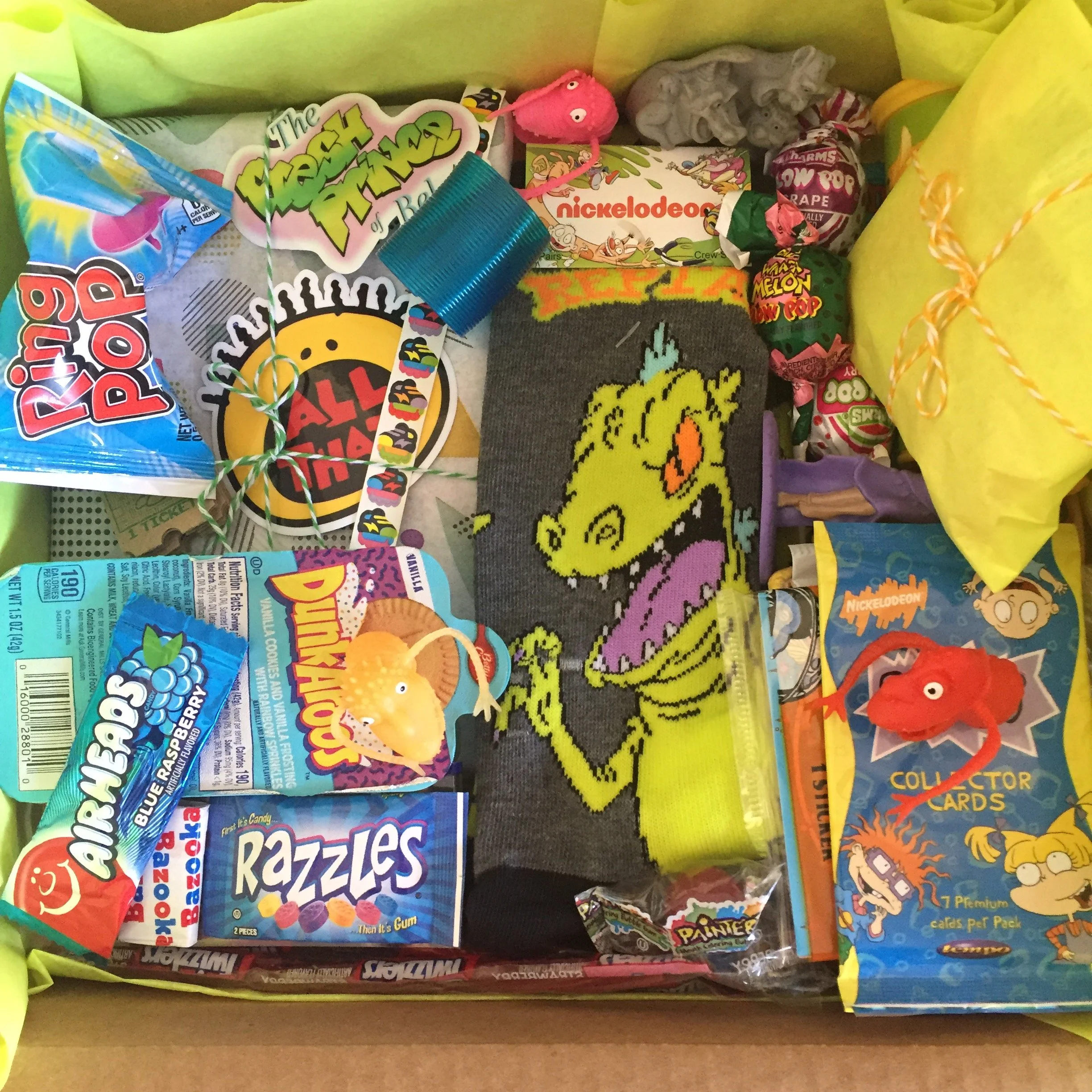 Image includes a variety of 90s themed products, include grey and green Reptar socks, yellow and red All That sticker, teal package of Dunkaroos, a little orange monster finger puppet, and candy including Ring Pop, Razzles, Blow Pops, and Airheads.