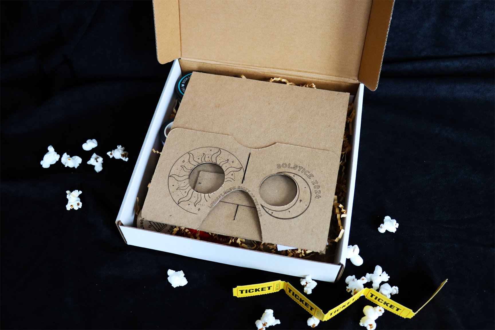 White cardboard gift box containing krinkle paper and a cardboard stereoscope viewer, folded flat. Yellow paper carnival tickets and popcorn are scattered around box.