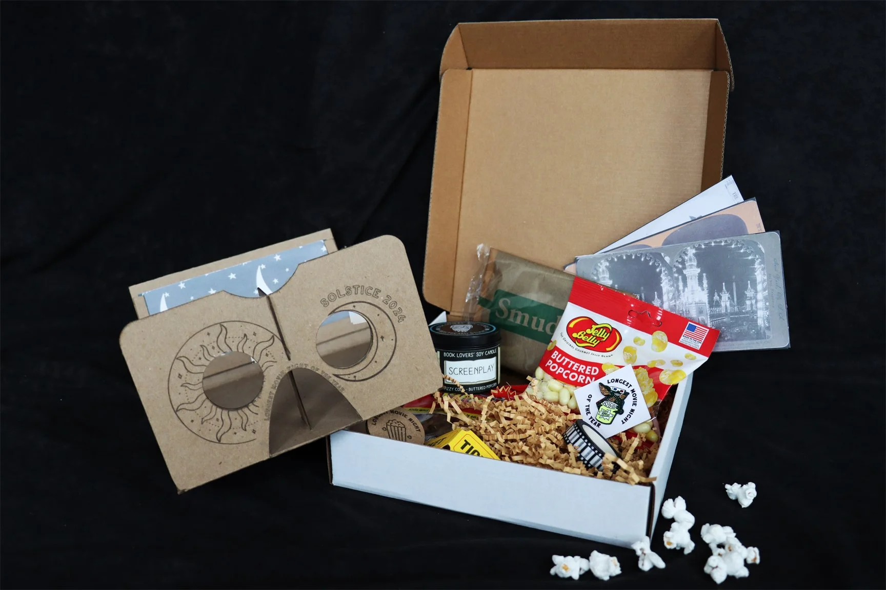 White cardboard gift box on black backdrop, containing buttered popcorn Jelly Belly candies, film strip patterned washi tape, scented candle labeled "Screenplay," a cardboard stereoscope viewer, kraft paper krinkles, and other items.
