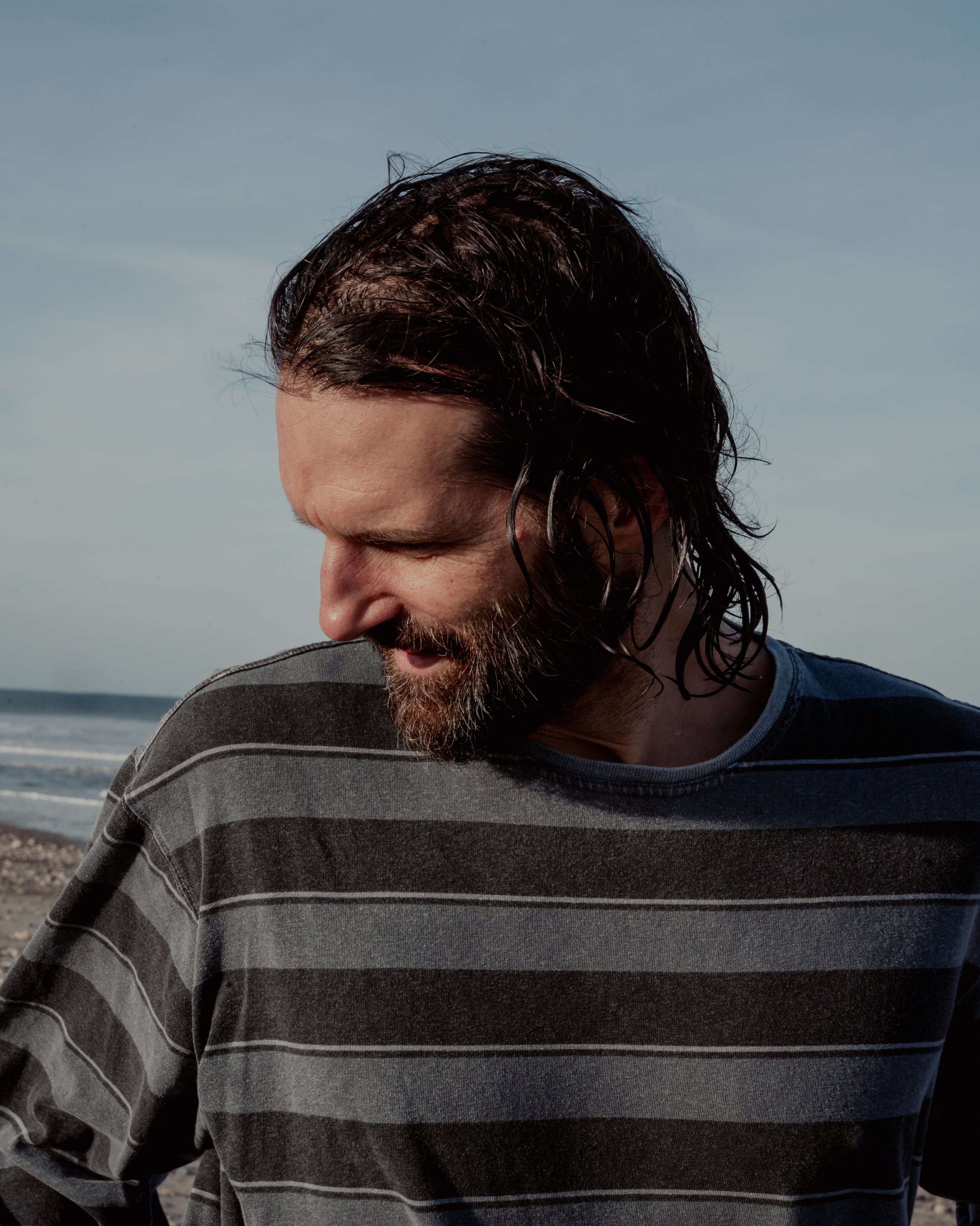 A man with long, dark, wet hair and a beard, wearing a striped gray and black shirt, standing on a beach with the ocean in the background and looking down.