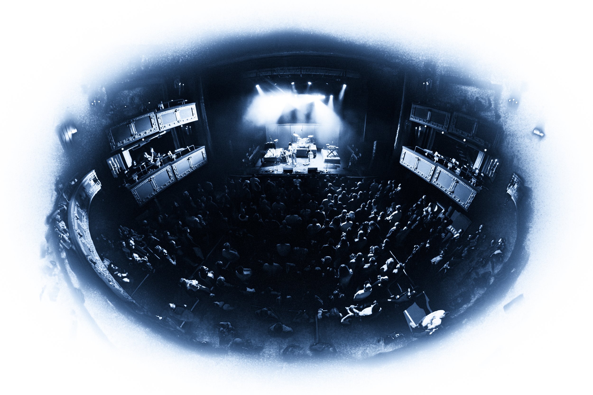 Crowd watching a live music concert on a stage with blue lighting, multiple performers, and balconies on either side of the theater.