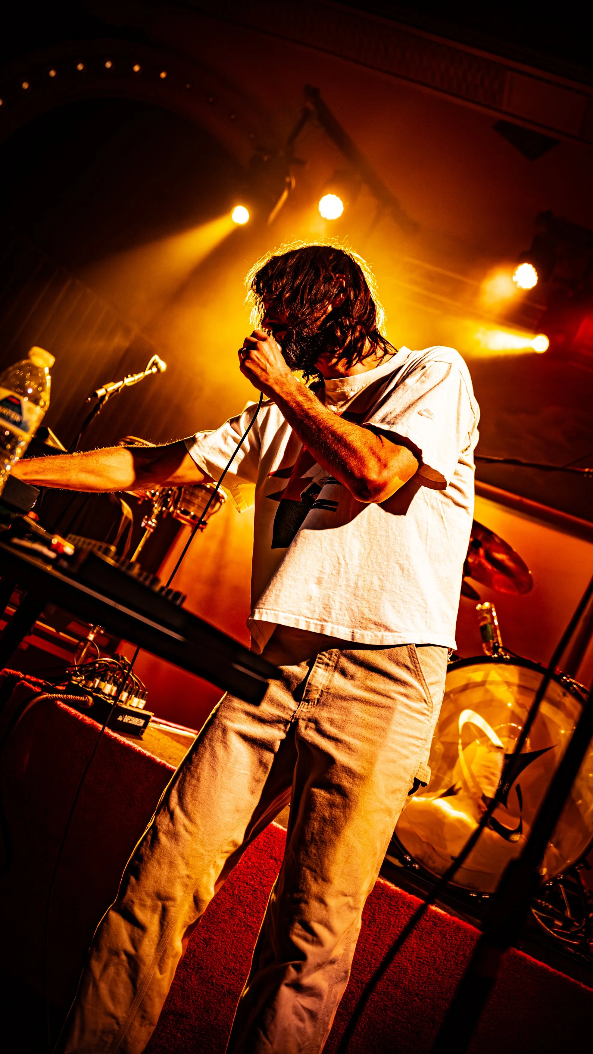 A male performer with long hair and a beard singing into a microphone on stage, surrounded by bright yellow and orange stage lights, with musical equipment including a drum set and a keyboard in the background.