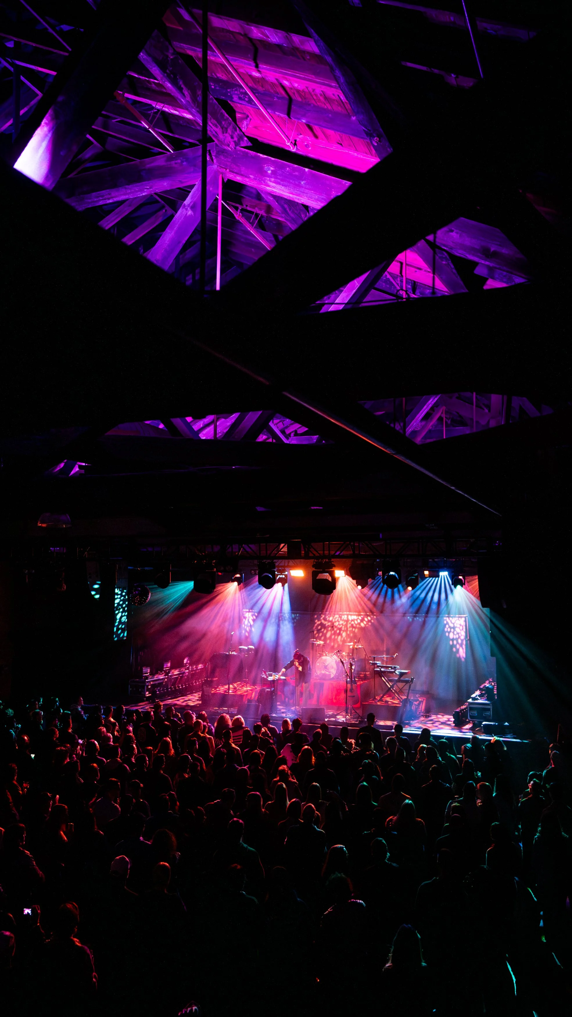 A concert with a large audience, stage lights illuminating a band, and a colorful light display on stage in a dark indoor venue.
