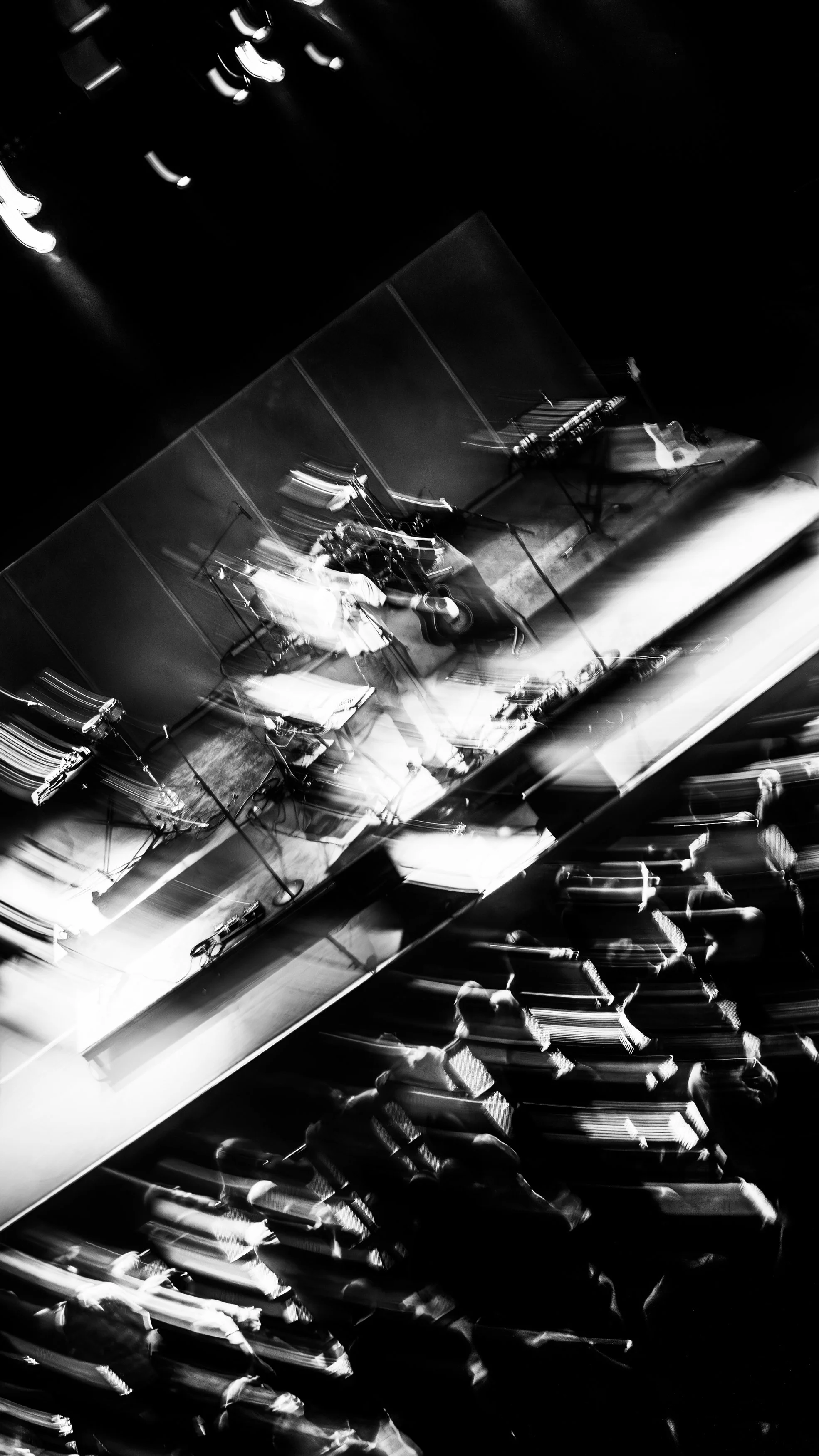 A black and white photo of a stage with musical instruments and microphones, and an audience sitting and standing in front of it, with some motion blur creating a dynamic effect.
