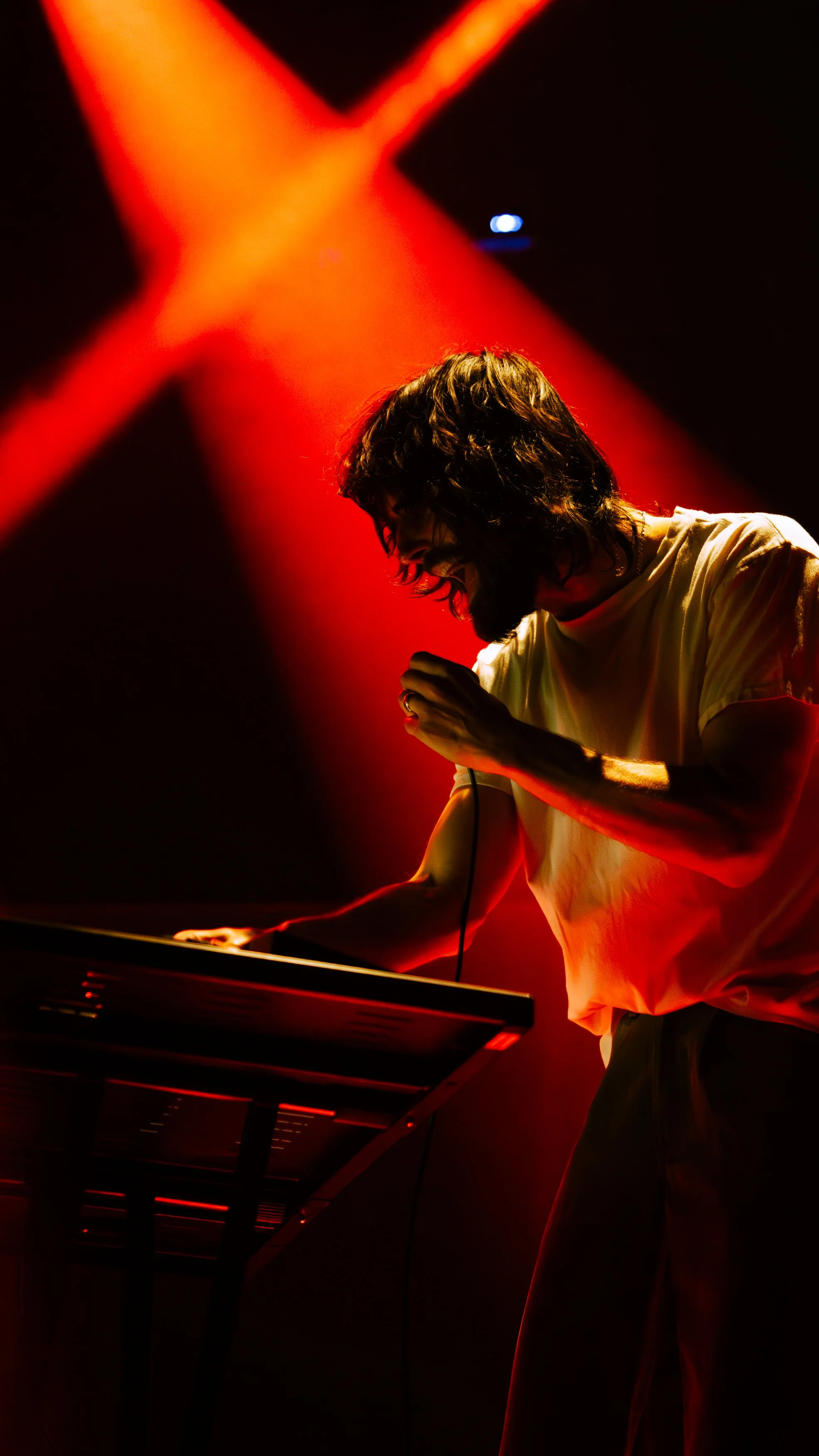 A man with glasses and long hair, illuminated by red stage lights, singing into a microphone while playing a keyboard on a music stage.