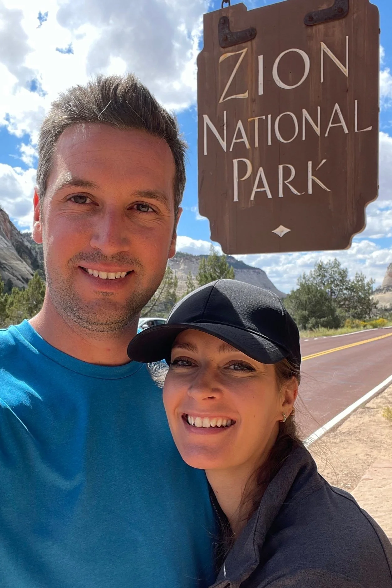 A smiling man and woman taking a selfie in front of a wooden sign that reads 'Zion National Park' with a mountainous landscape and partly cloudy sky in the background.
