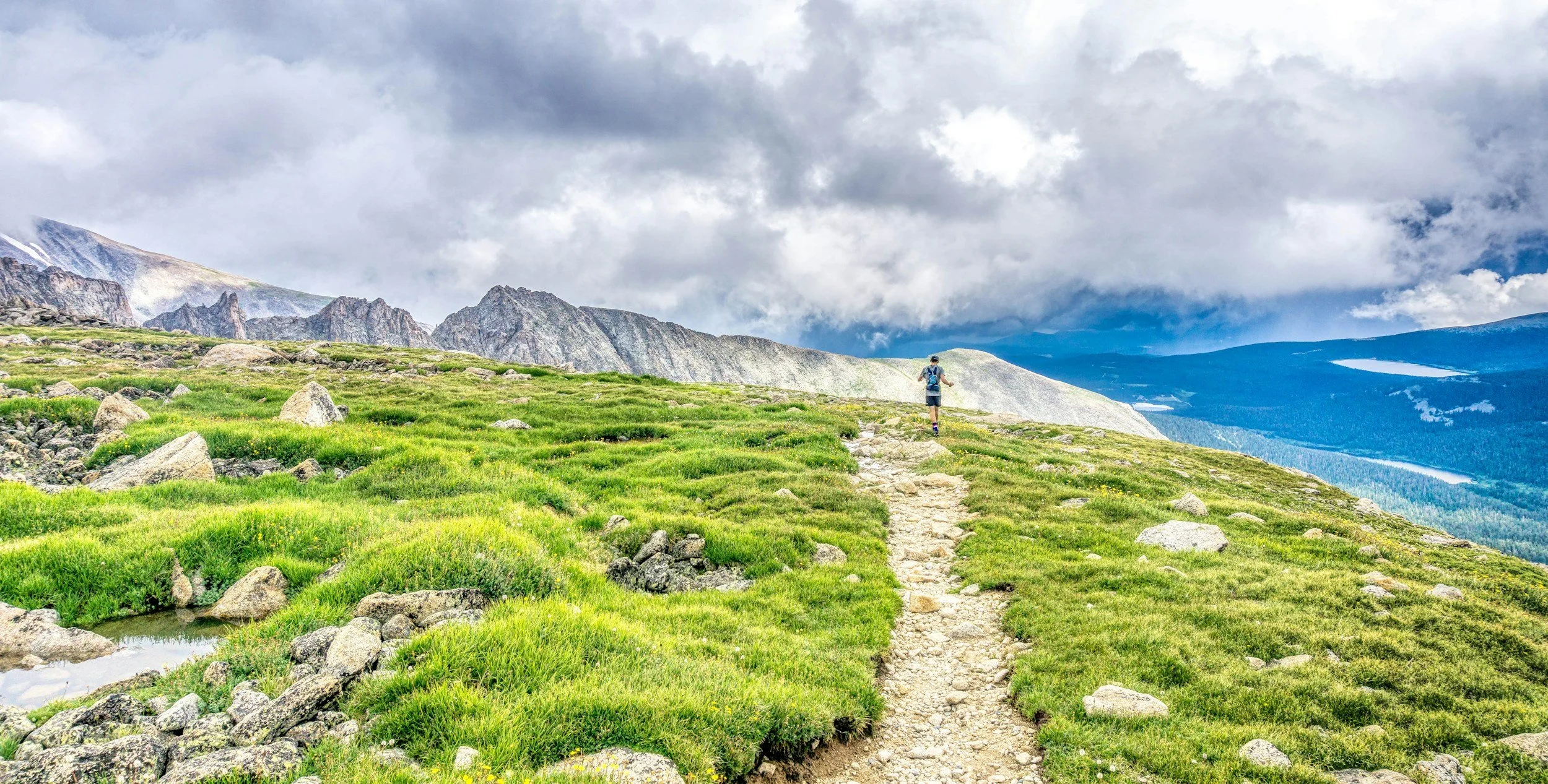 A person hiking on a narrow dirt trail through a green mountain meadow with rocky terrain, surrounded by distant mountains and cloudy sky.