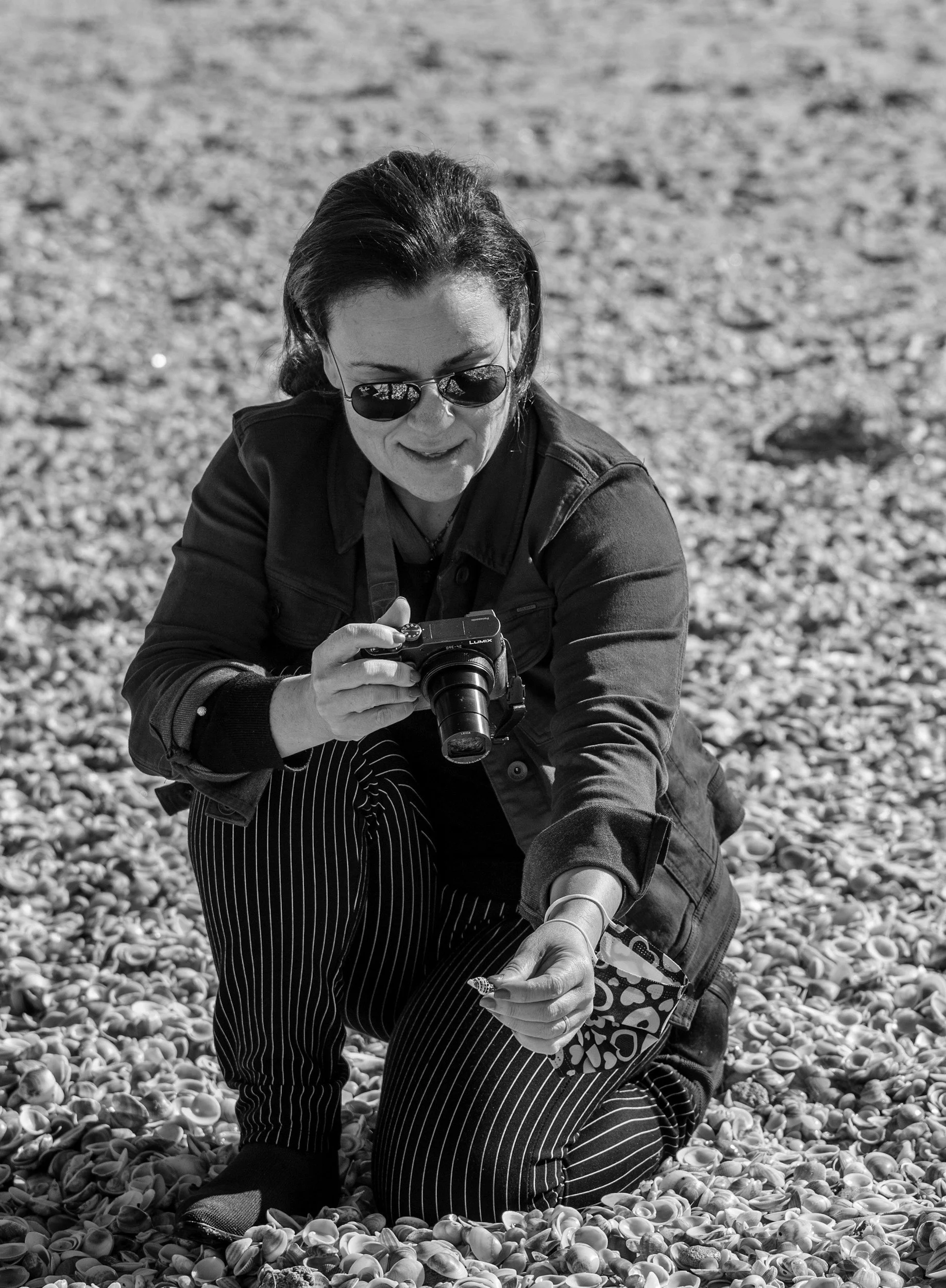 A woman crouching on a shell-covered beach, wearing sunglasses and striped pants, holding a camera.