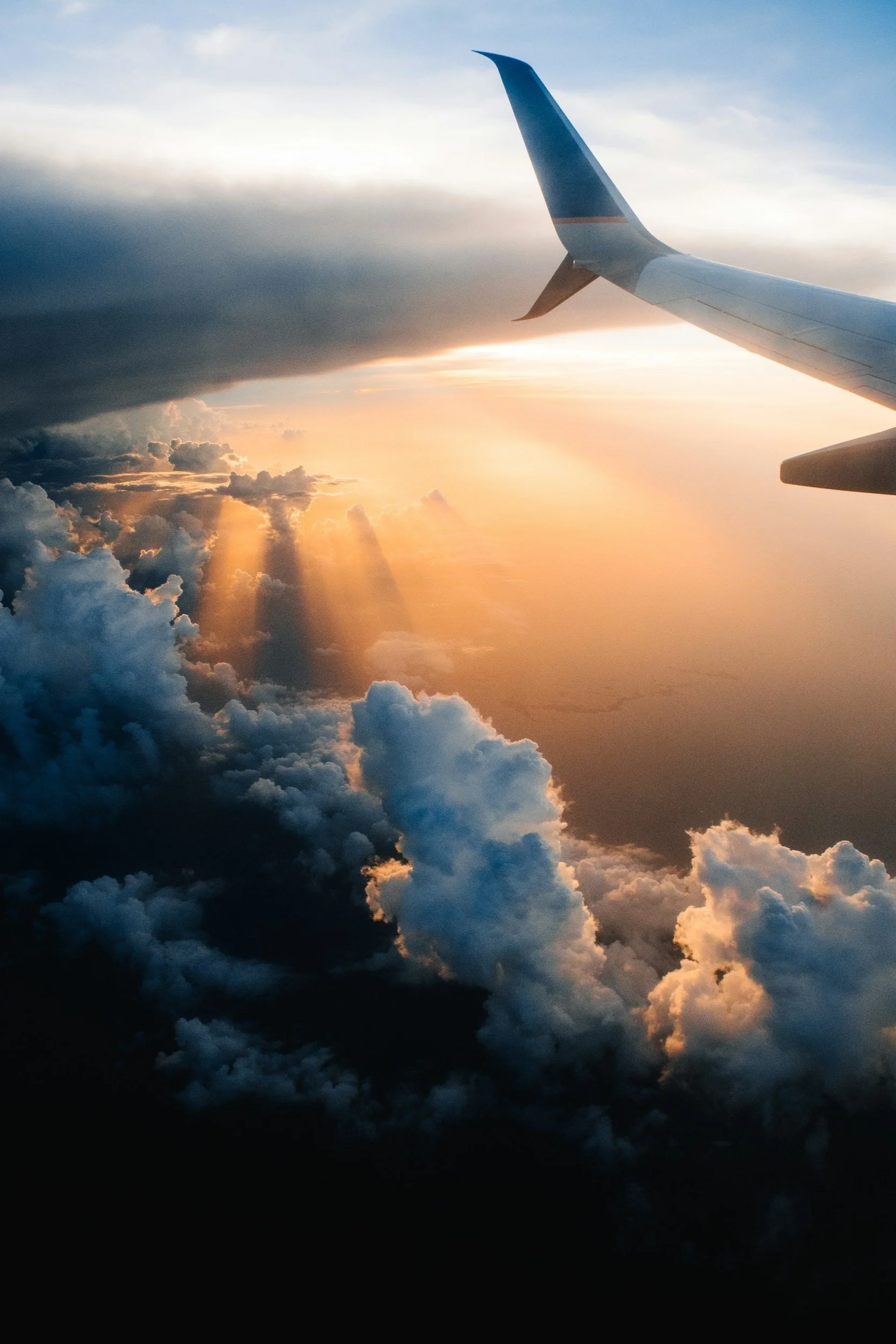 Photo taken from an airplane window showing the airplane wing in the sky, with sunlight breaking through clouds during sunset or sunrise.