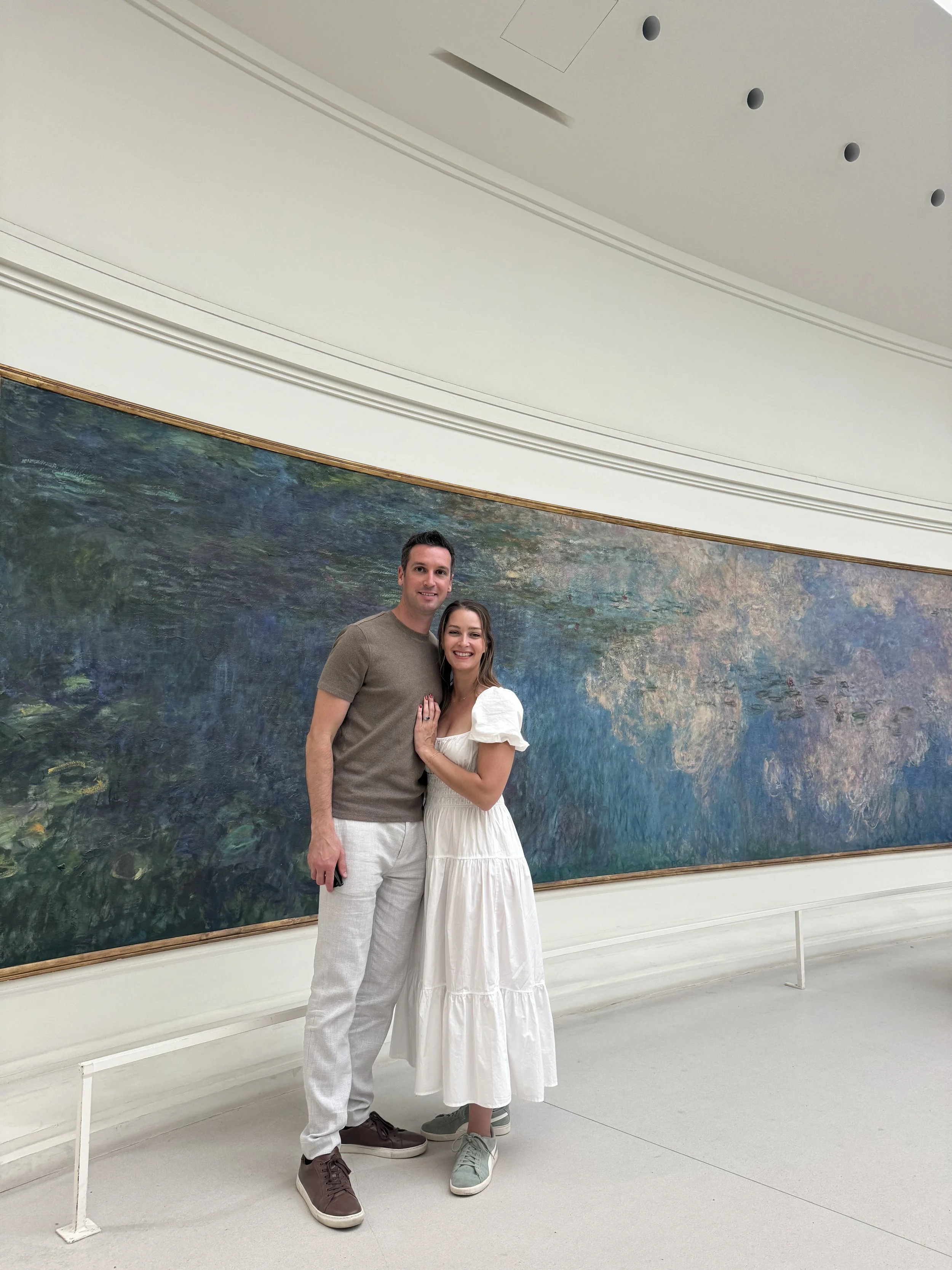 A man and woman standing in front of an abstract landscape painting in an art gallery, smiling at the camera.
